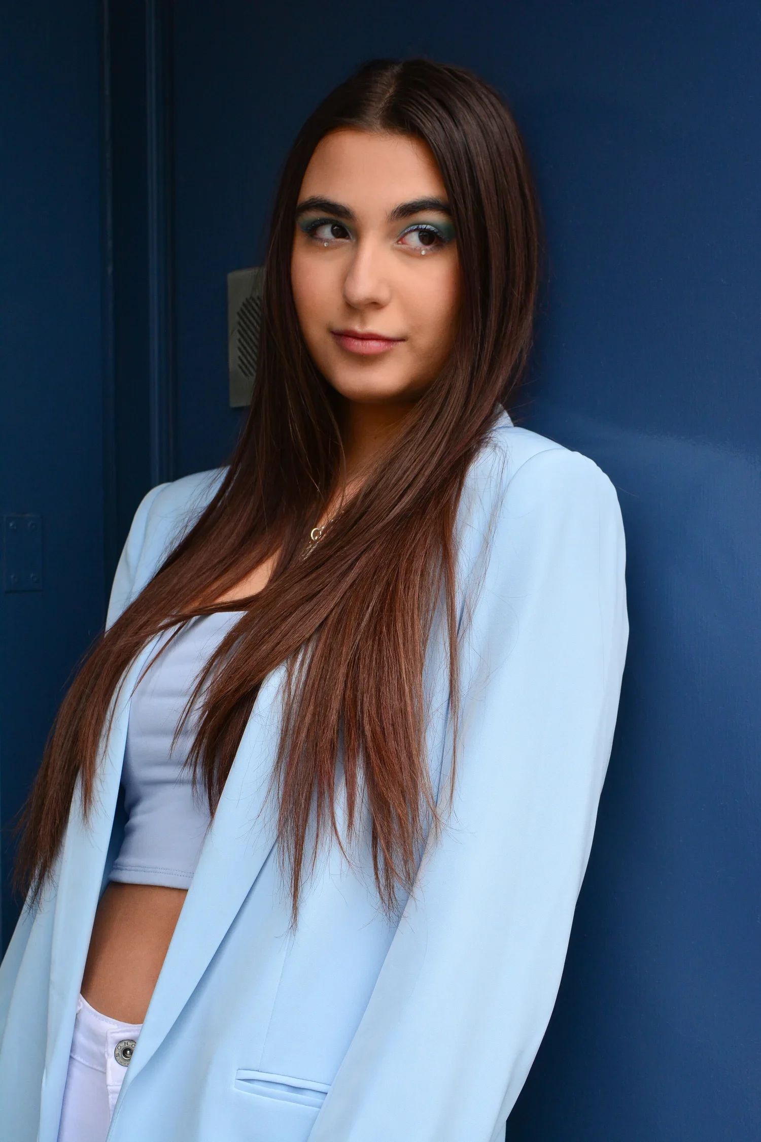 Young woman with long brown hair and blue eyeshadow standing against a dark blue wall, wearing a light blue blazer and crop top.
