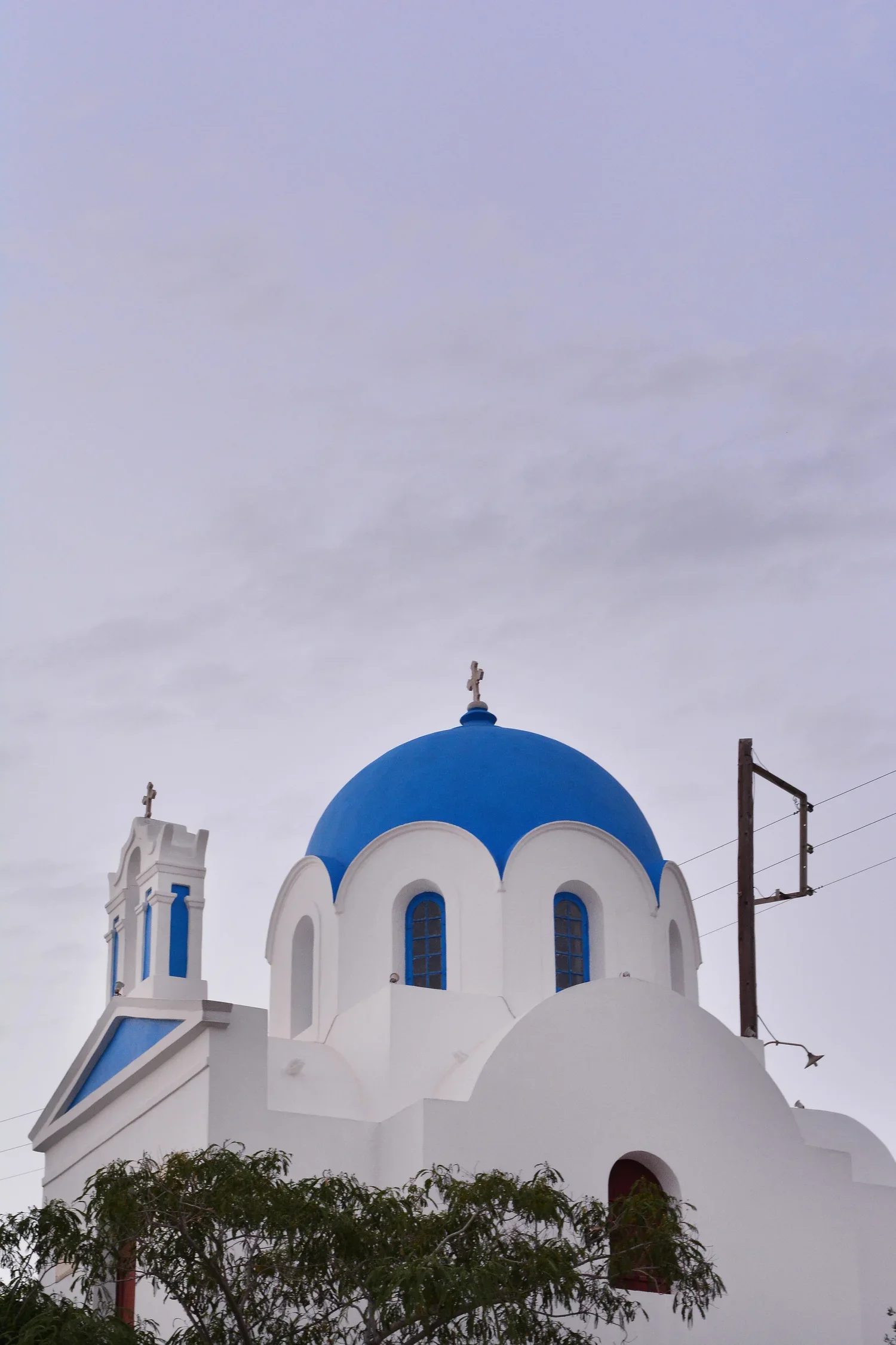 A white Greek Orthodox church with a blue dome and crosses on top, located under a gray sky, with trees in the foreground.