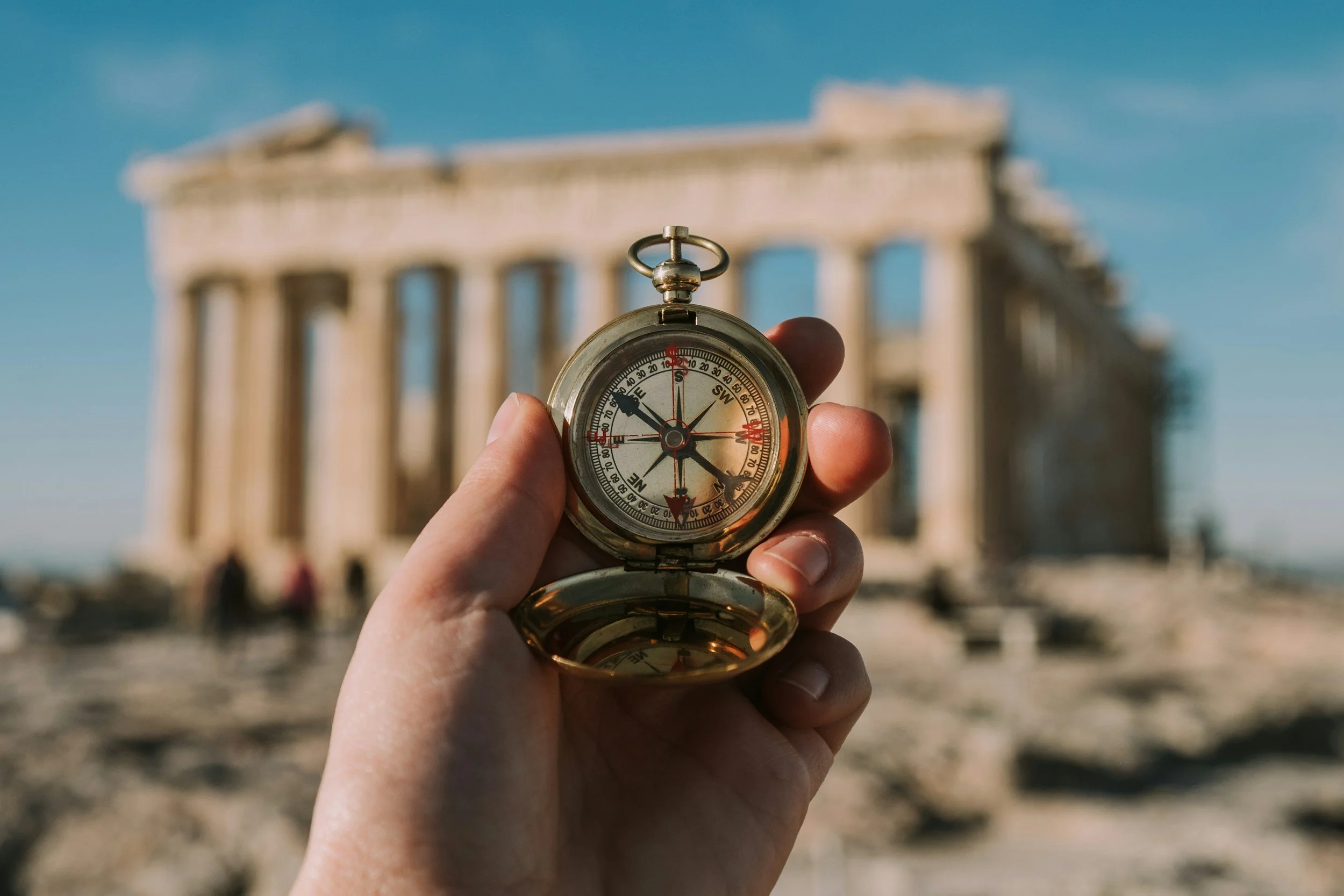 A hand holding a compass in front of the ancient Parthenon temple in Greece.