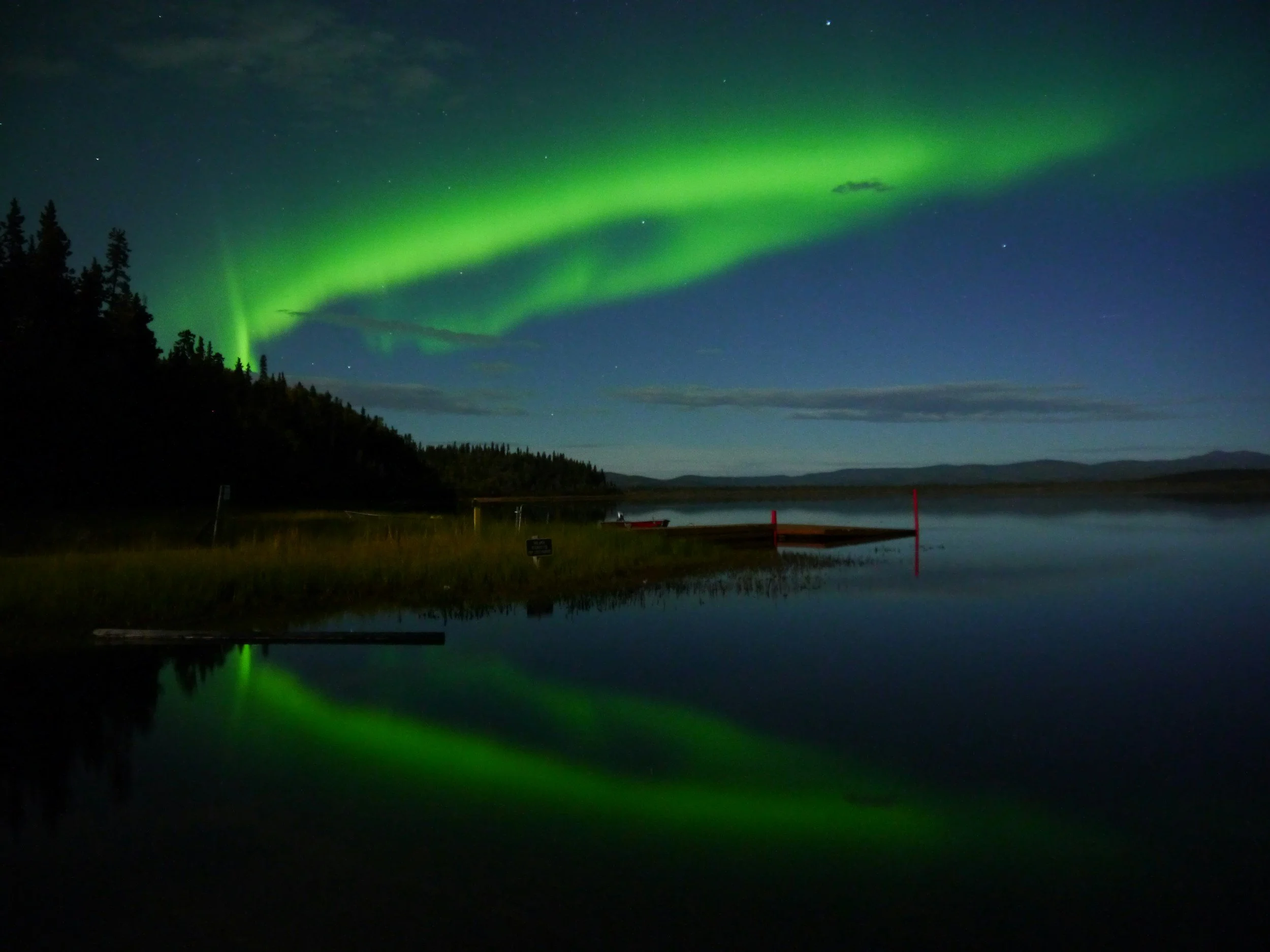 Northern lights over a lake at night with reflections, trees on the shoreline, and a partly cloudy sky.