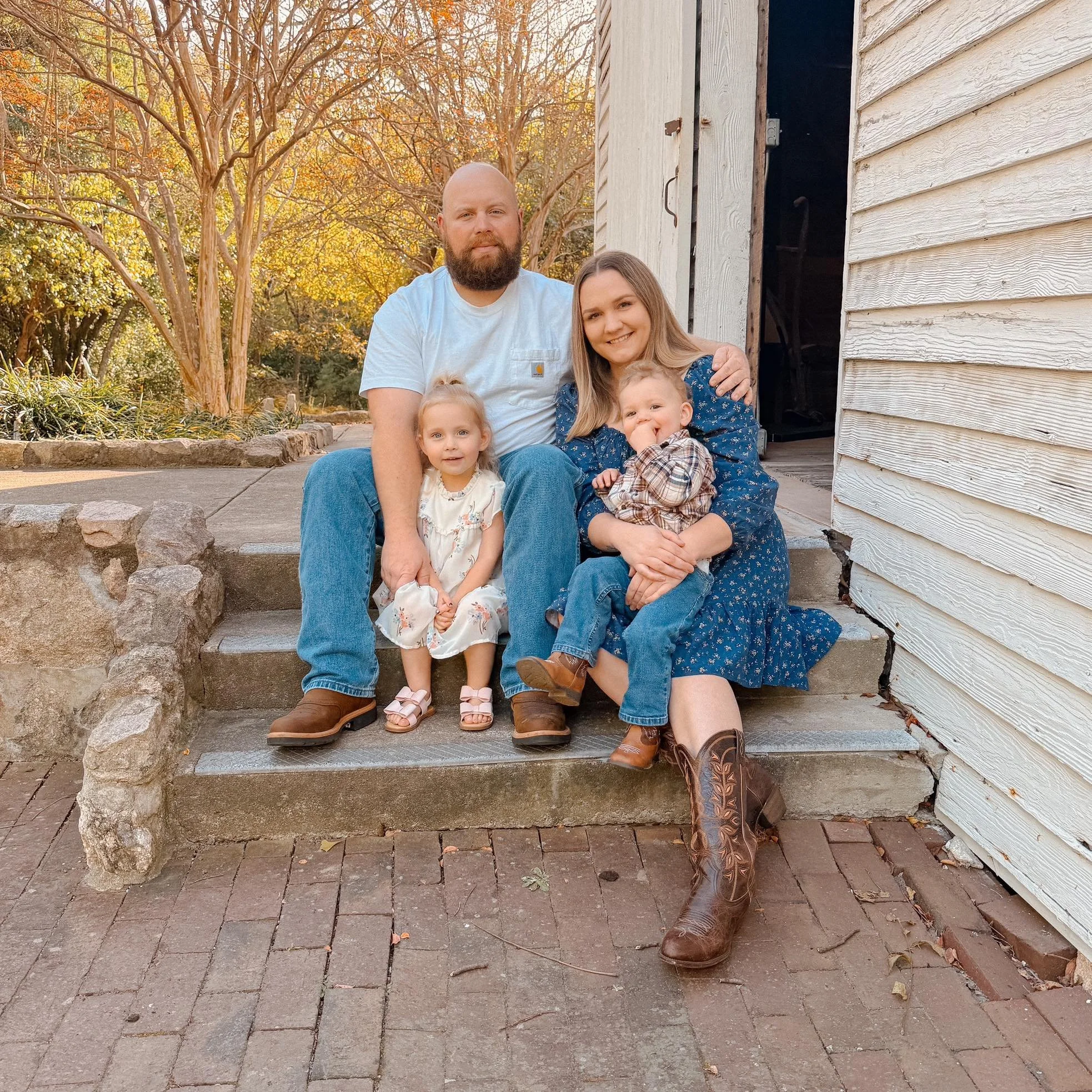 A family of four sitting on outdoor steps in front of a white wooden building with trees in the background. The dad has a beard and wears a white t-shirt and jeans. The mom has long hair and wears a blue dress with floral patterns. The young girl is in a white dress and pink sandals. The young boy is in a plaid shirt, jeans, and brown boots. The family is smiling at the camera during autumn.