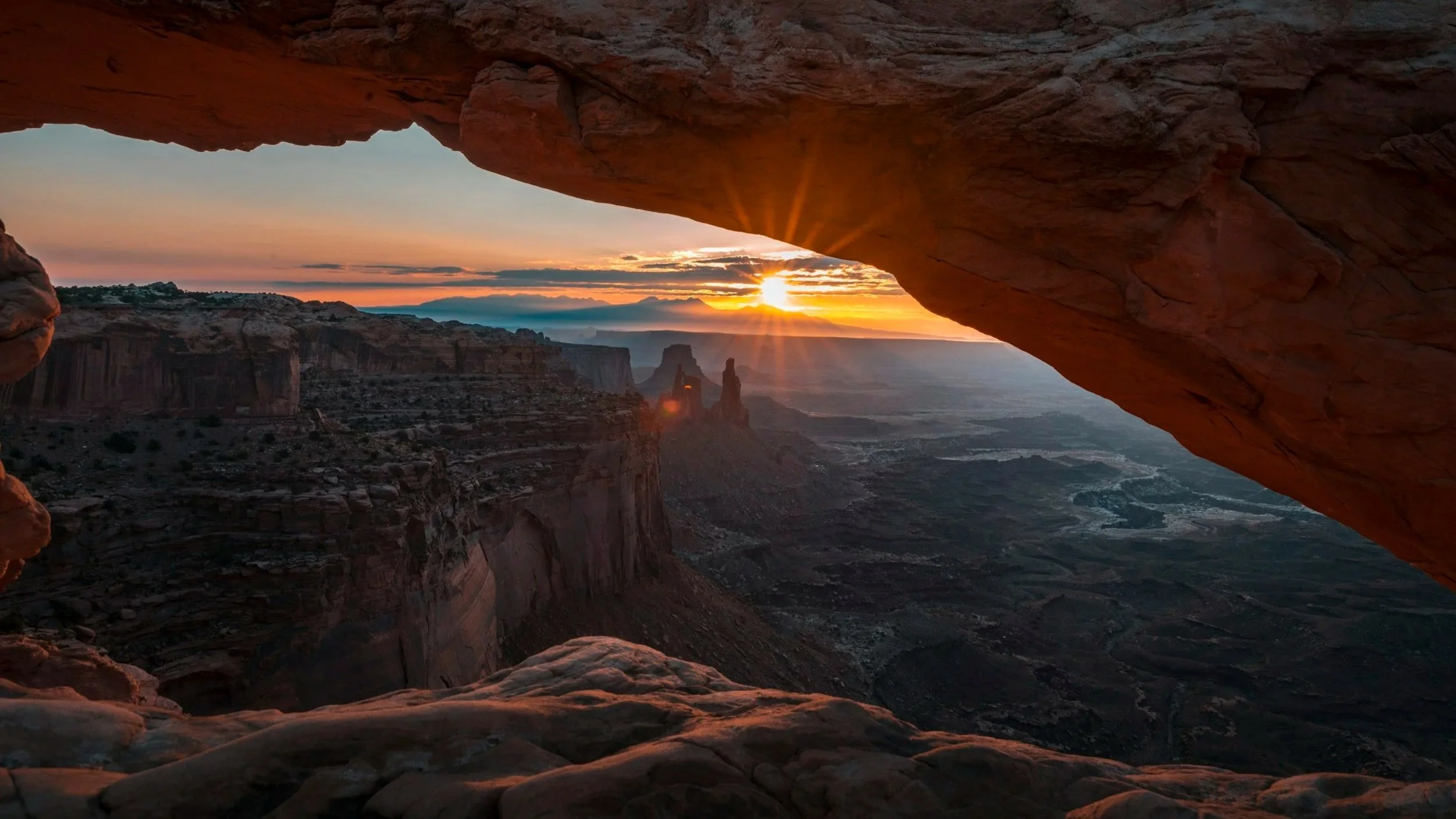Sunset view through a natural rock arch overlooking a canyon with distant rock formations and a vibrant sky with clouds.