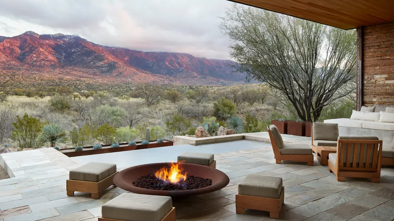 A modern outdoor patio with seating around a firepit, overlooking a desert landscape with mountains in the background.
