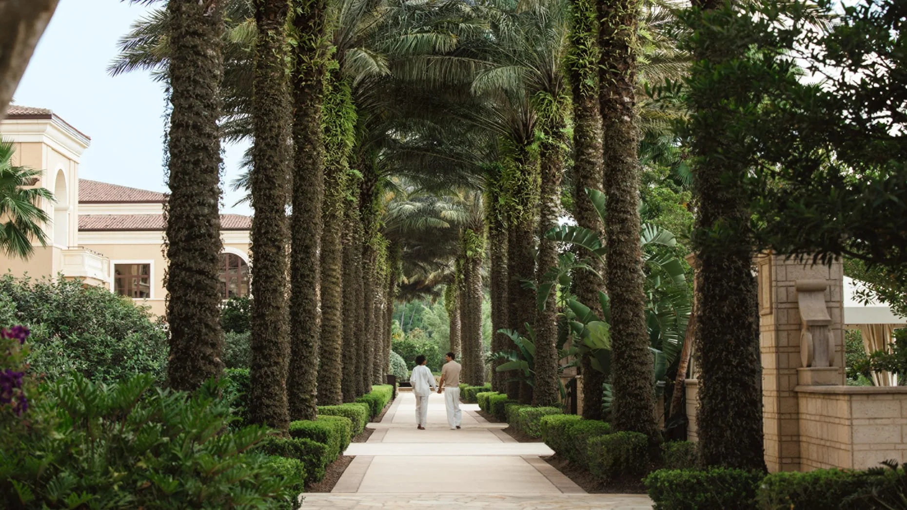 A couple walking hand-in-hand along a landscaped pathway flanked by tall palm trees and green bushes on a sunny day.