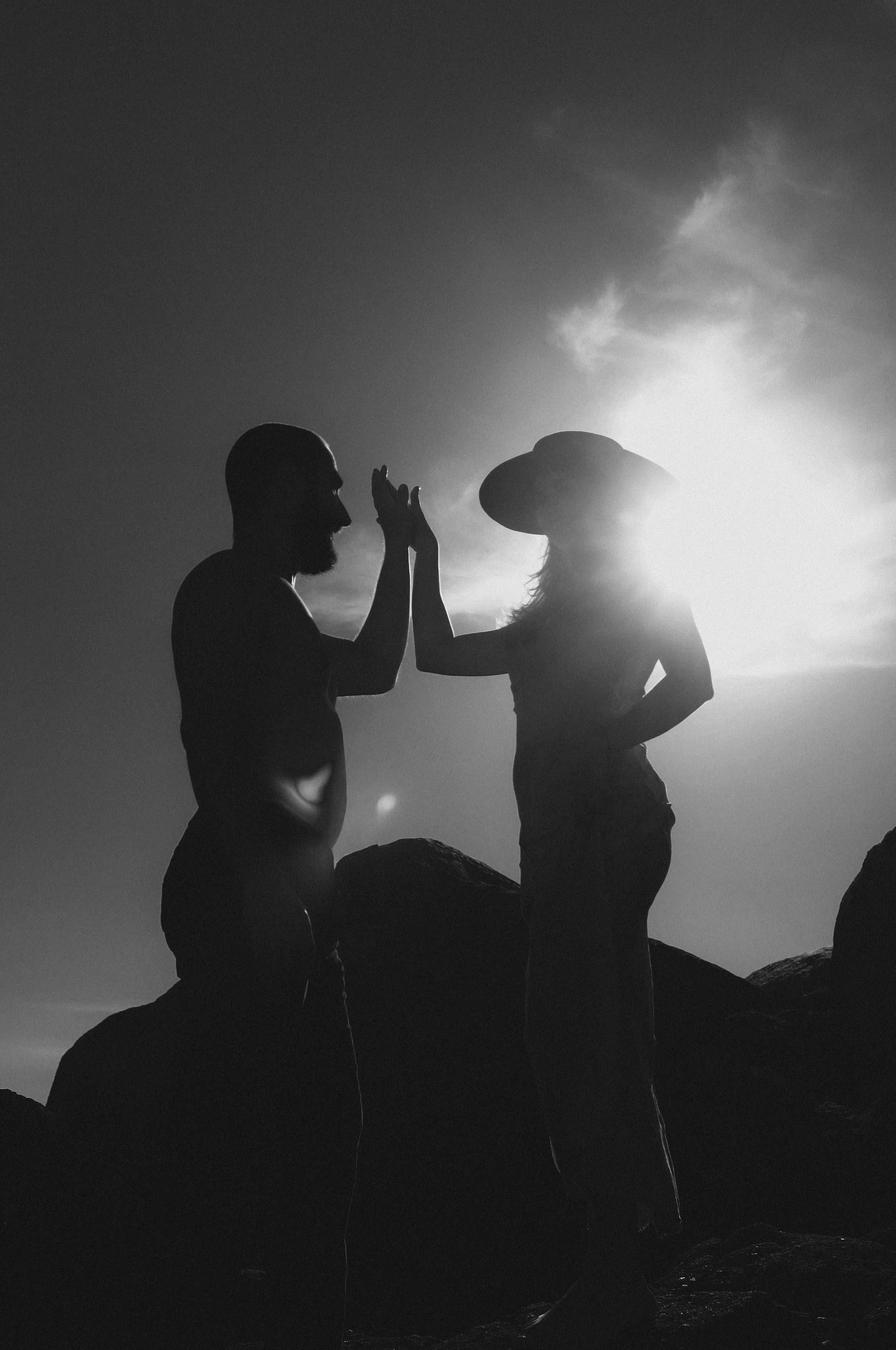 Silhouettes of a man and woman with foreheads touching and hands raised, facing each other on a rocky landscape with the sun setting behind them.