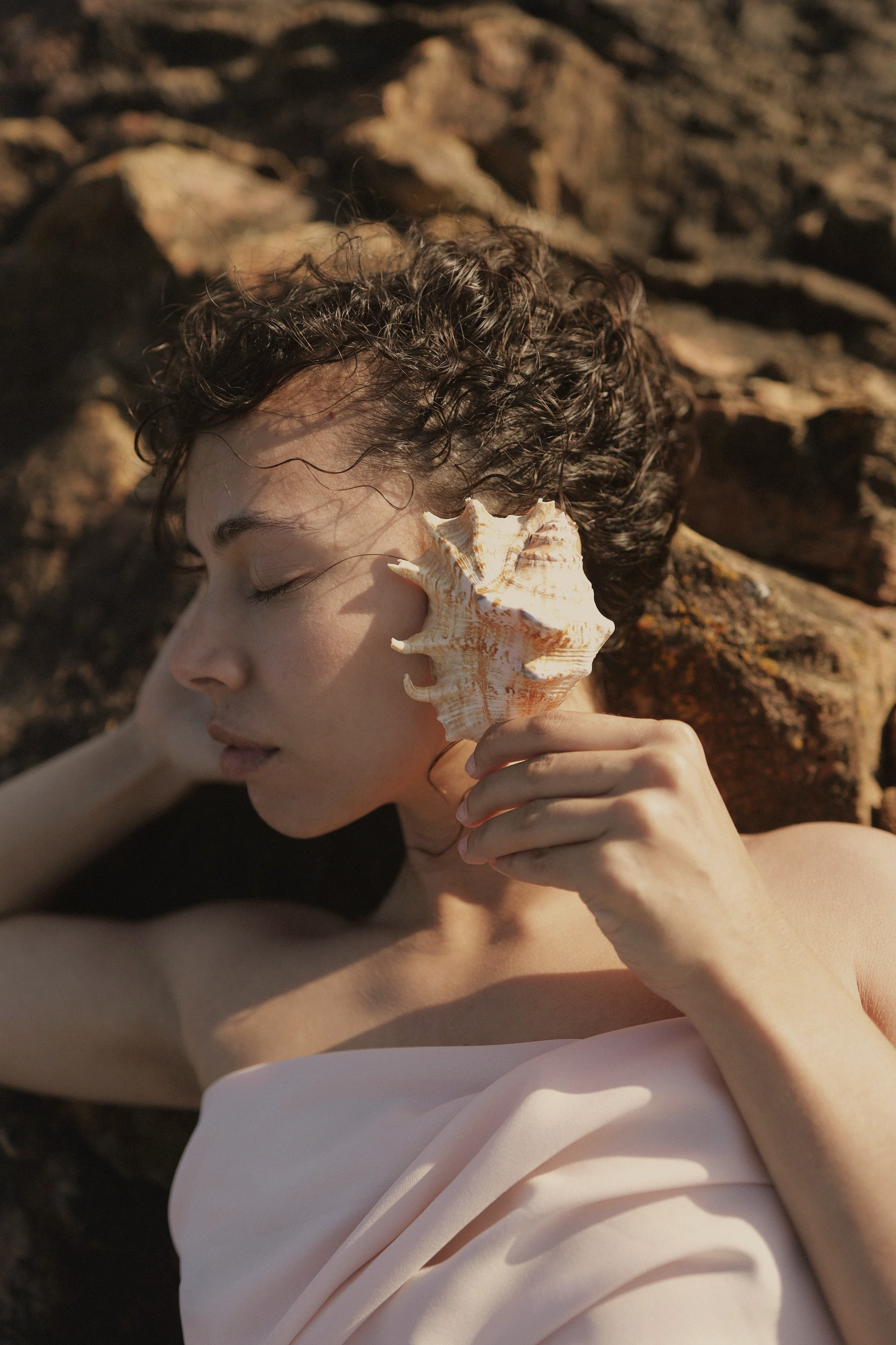 A woman with curly dark hair and light skin is lying on rocks with her eyes closed, holding a large seashell to her ear, wearing a white garment.