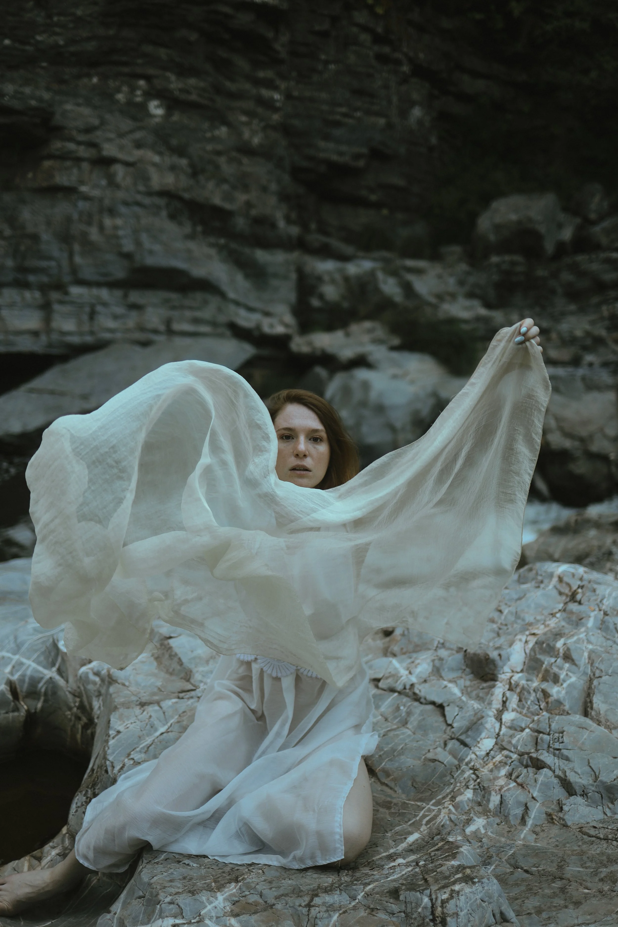 A woman with long brown hair and light skin, dressed in white, standing among rocks and holding a flowing white fabric, with a large rock formation in the background.