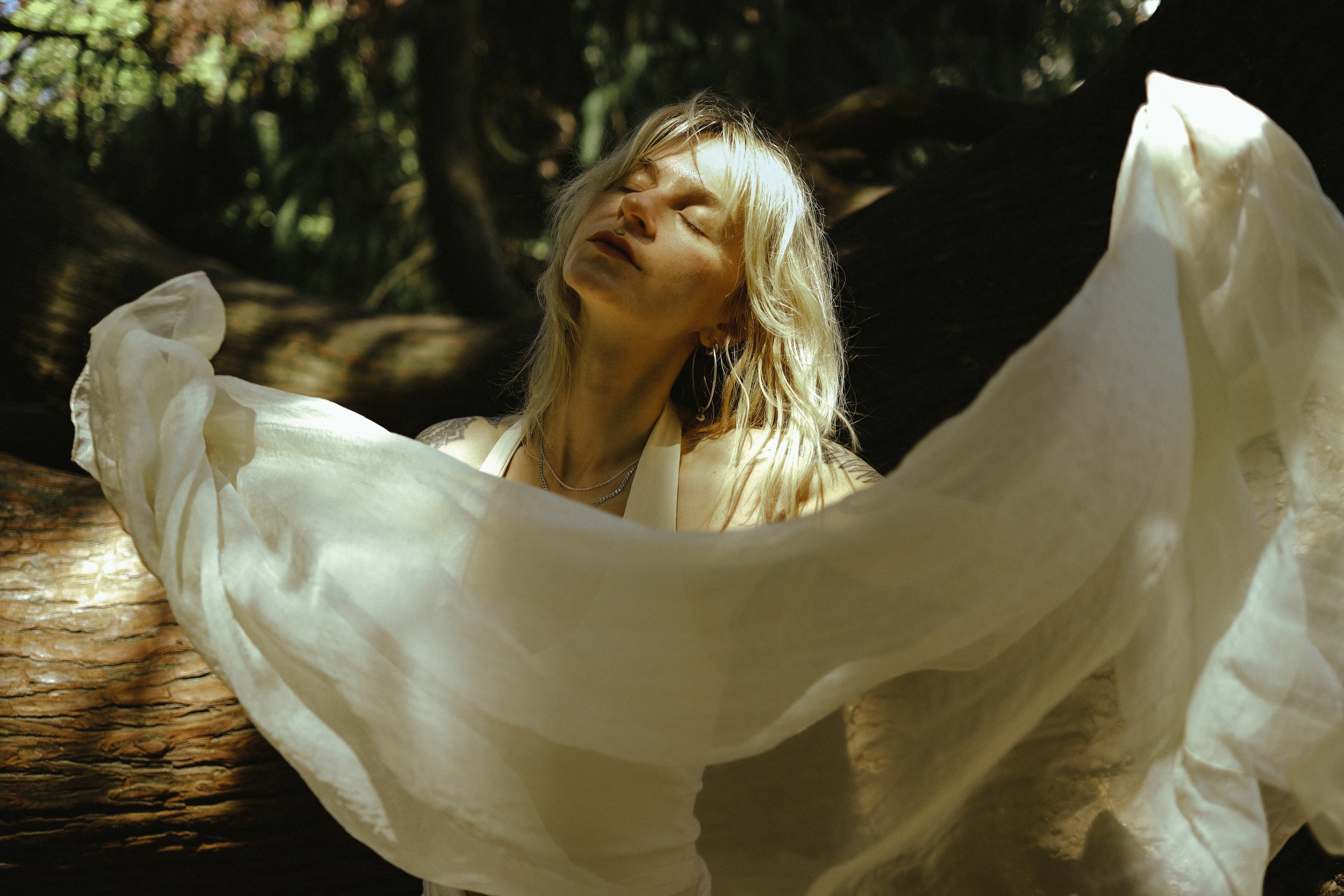 A person with blonde hair, closed eyes, and a serene expression, is outdoors in a wooded area, surrounded by tree branches and leaves, with sunlight filtering through the trees.