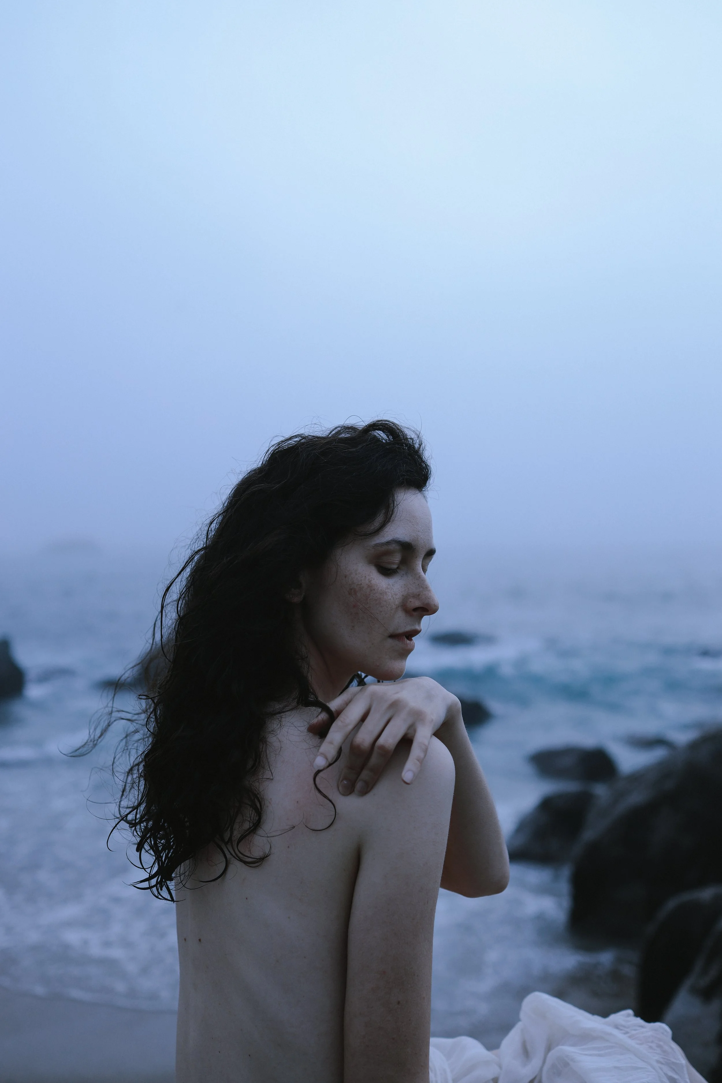 A woman with long, dark, curly hair and fair skin stands topless on a rocky beach at dusk with her eyes closed and one hand resting on her shoulder, facing toward the right side of the image.