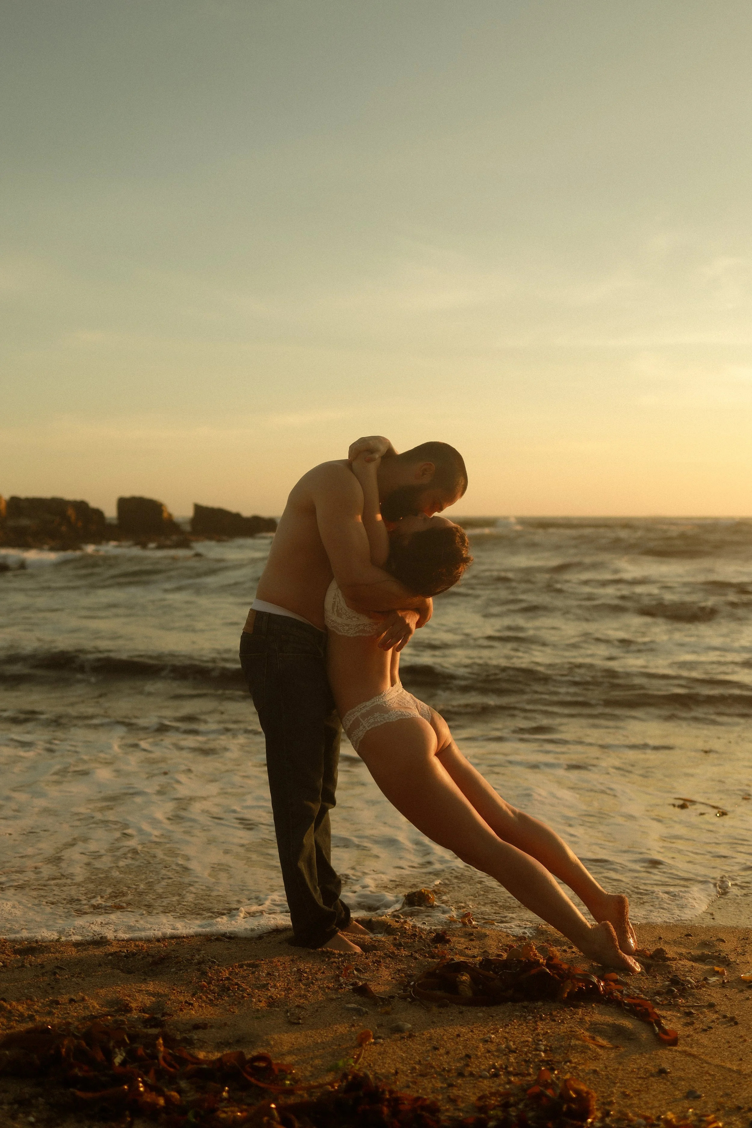 A couple dancing closely on a beach during sunset, with the man embracing the woman and the woman leaning back, both dressed in summer clothing, with rocks and seaweed on the sand and the ocean in the background.