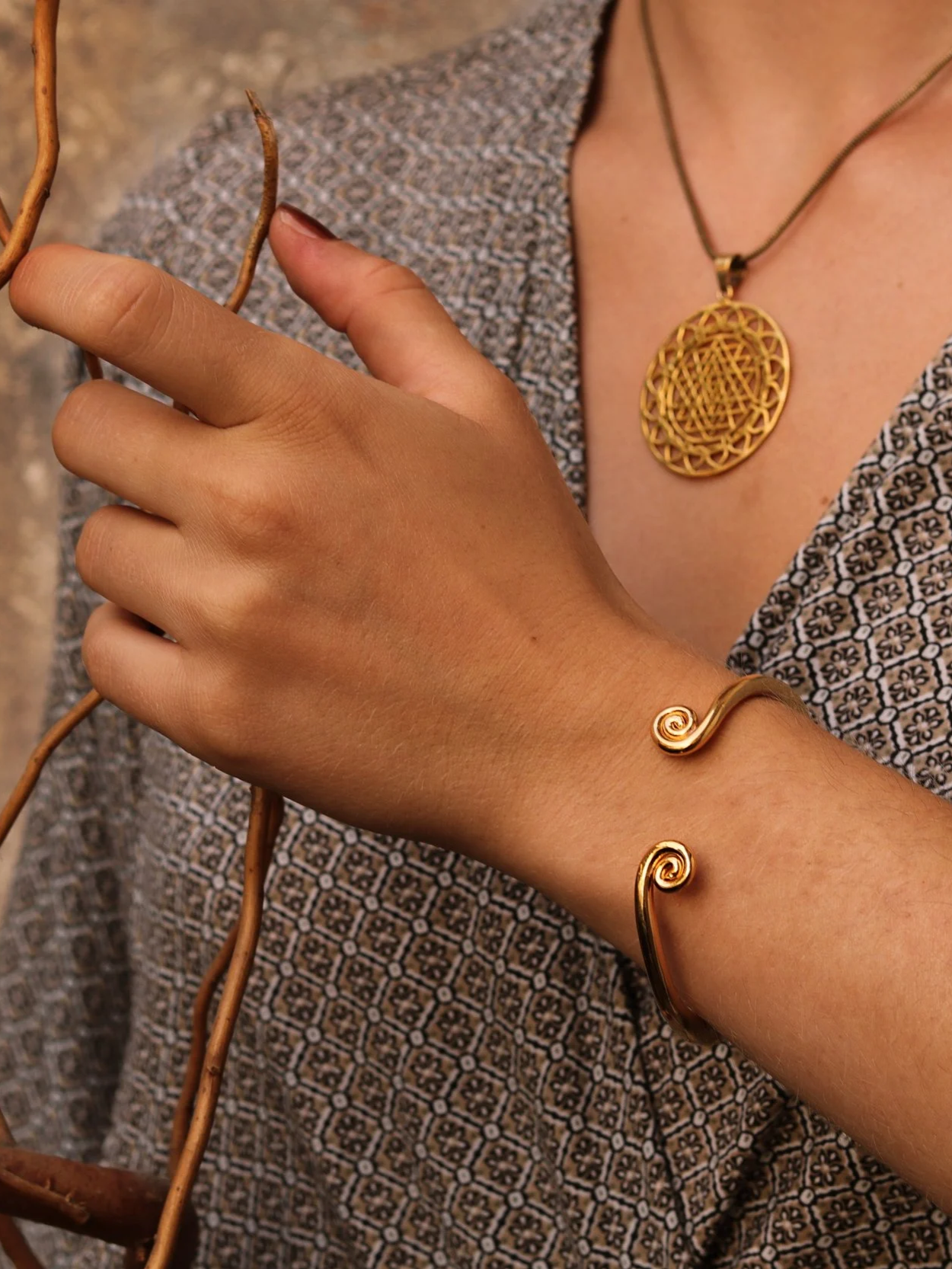 Close-up of a woman's hand and wrist adorned with gold spiral bracelets, holding dried plant stems, wearing a patterned dress, and a large gold pendant necklace.