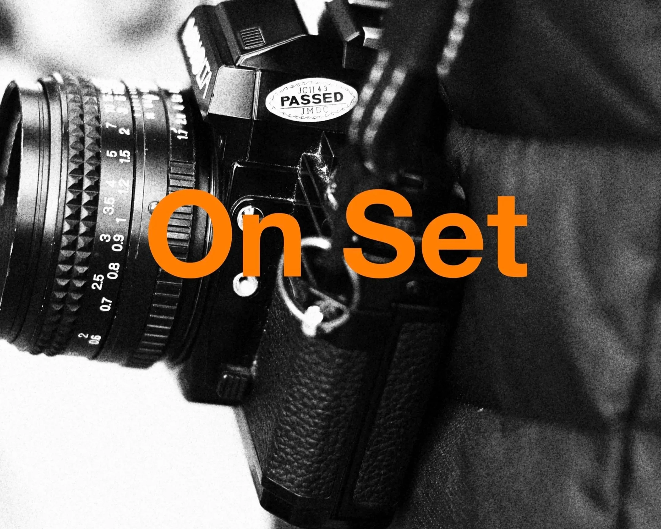 Black and white close-up photo of a camera lens with focus and zoom rings, bearing an orange overlay with the words 'On Set' in large font.