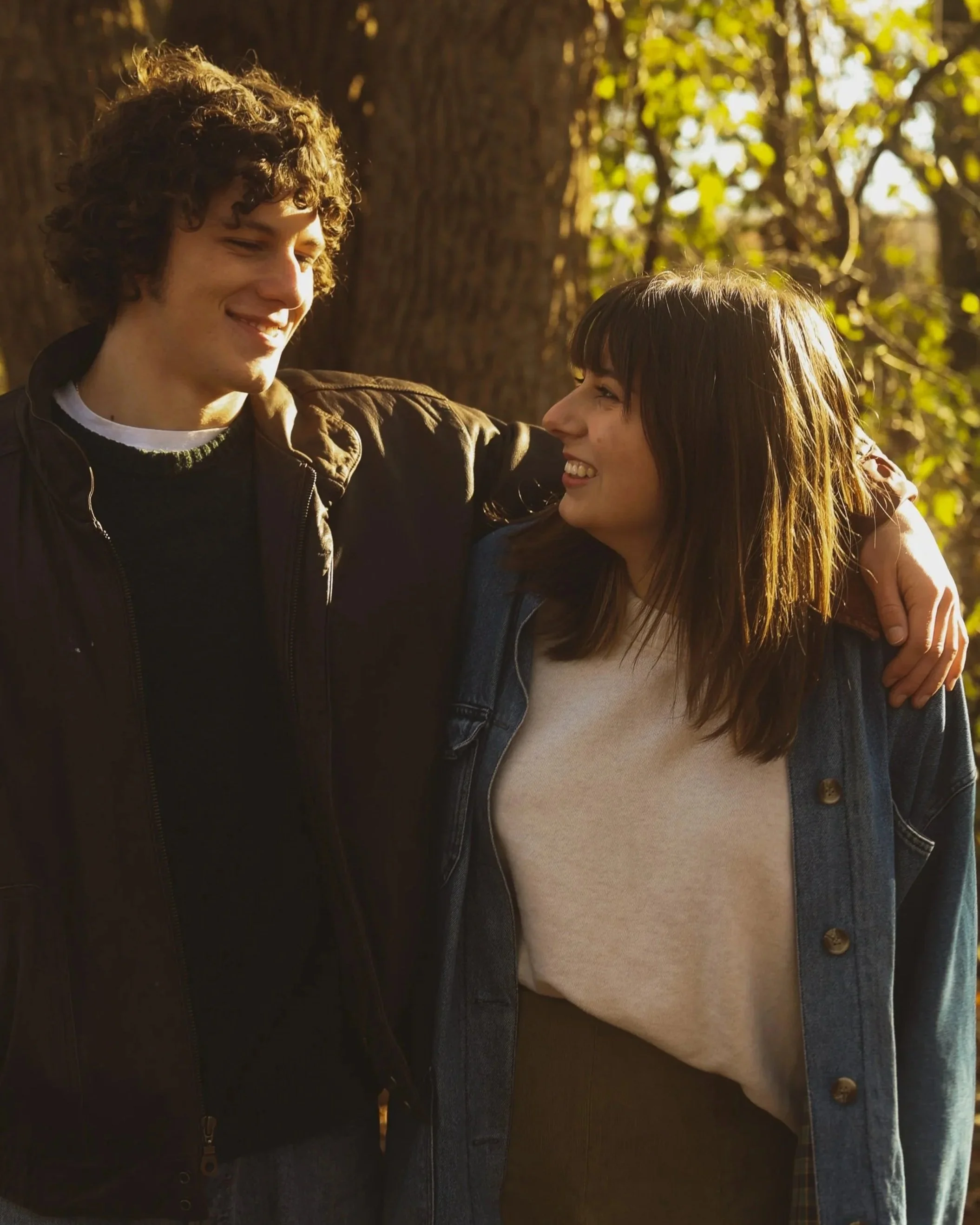 Two young people, a man and a woman, smiling at each other outdoors during a sunny day. The man has curly hair and is wearing a dark jacket, while the woman has straight hair with bangs and is wearing a light-colored shirt and a denim jacket. There a