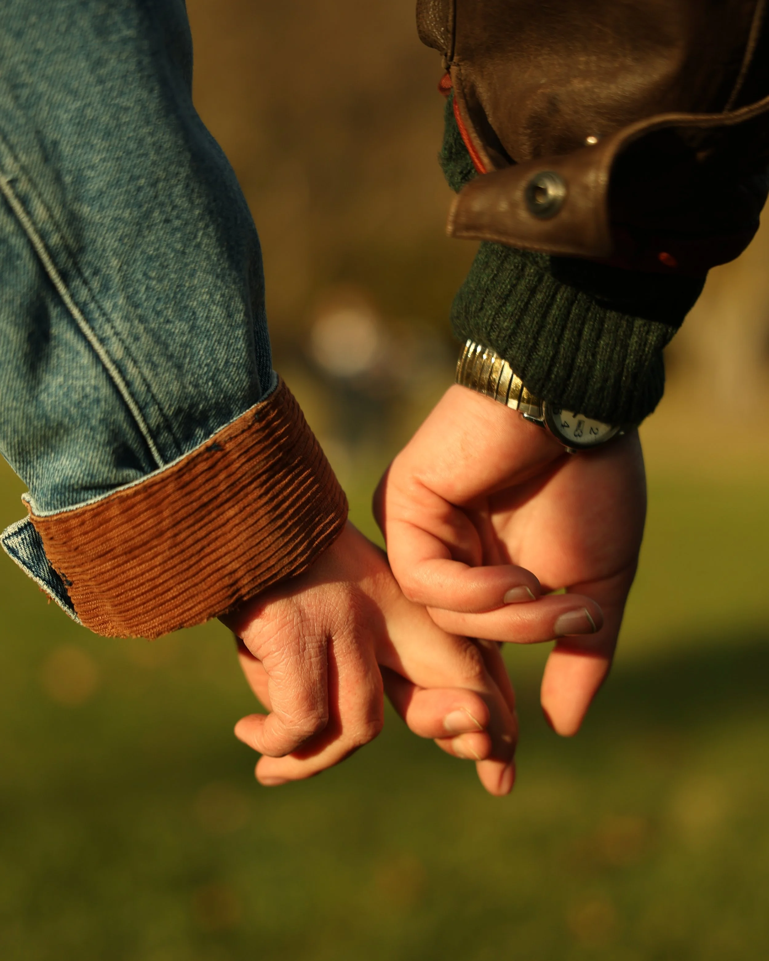Close-up of two hands holding each other, with one person wearing a watch and the other wearing a sweater and denim jacket, against a blurred natural background.