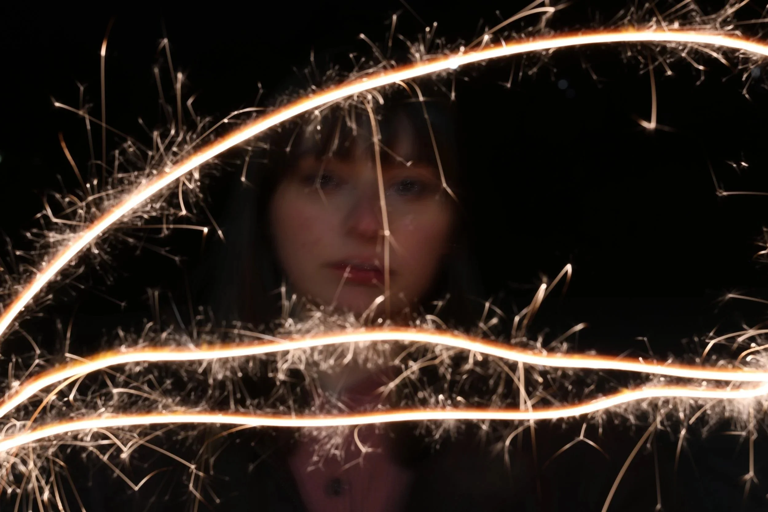 A person's face partially visible behind glowing sparkler trails on a dark background.