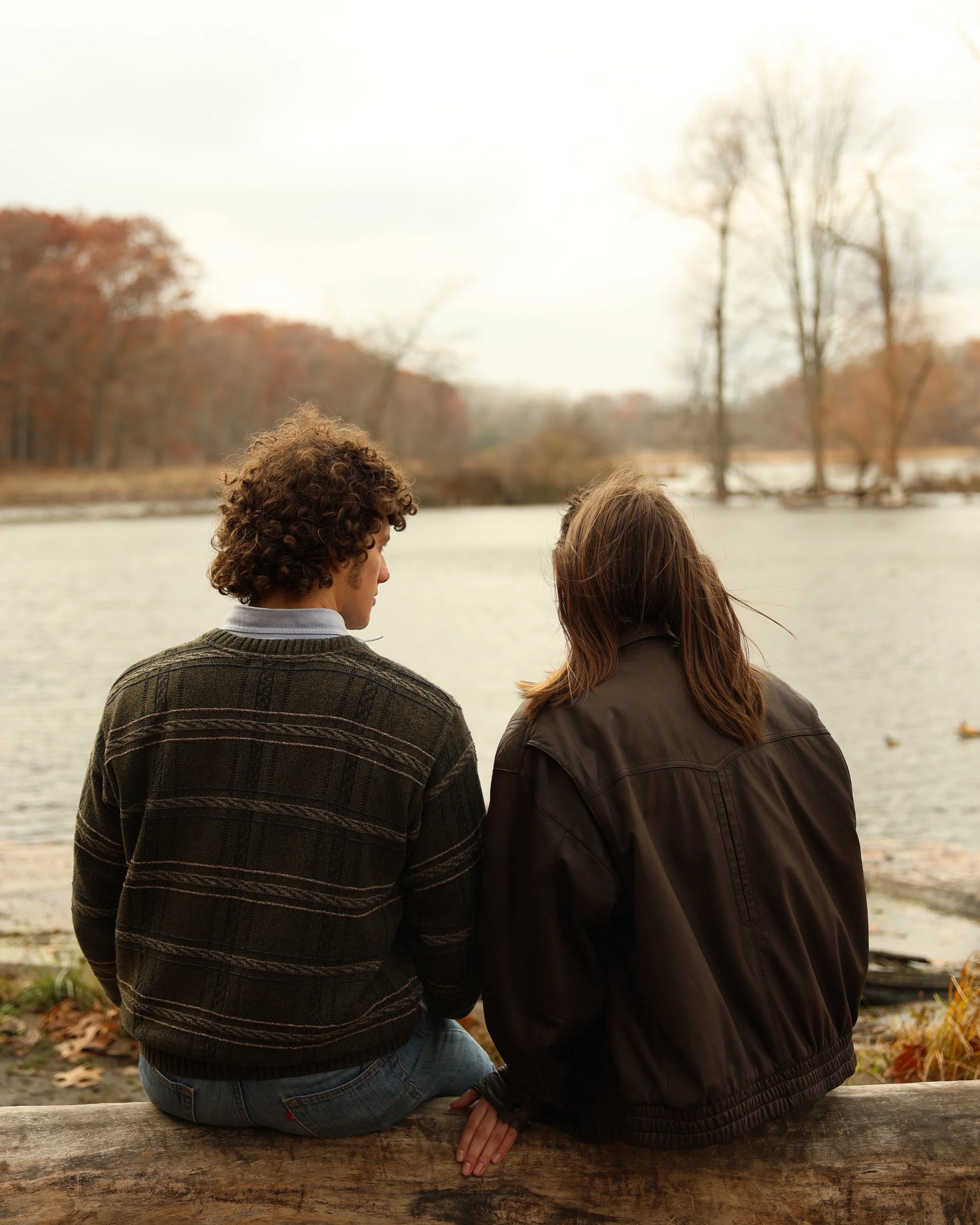 Two people are sitting on a bench near a river, with their backs to the camera. The person on the left has curly hair and is wearing a striped sweater and jeans. The person on the right has straight hair and is wearing a dark jacket. The background f