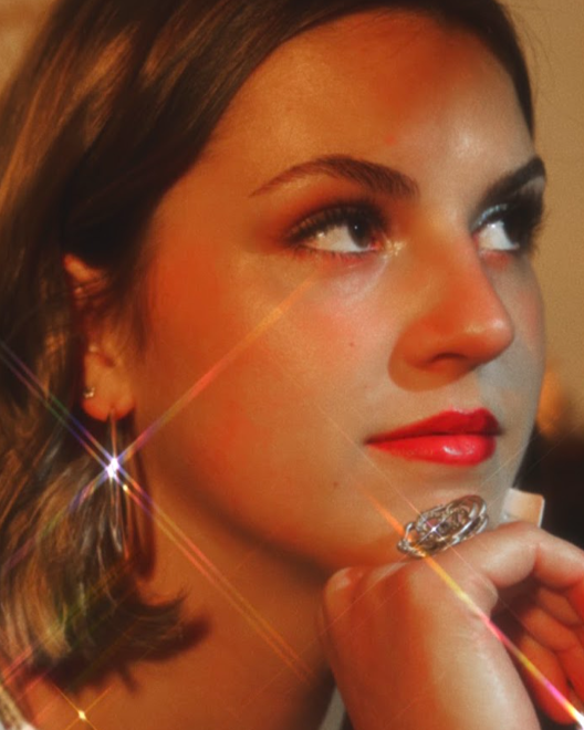 Close-up of a young woman with short brown hair, wearing earrings and a ring, crossing her arms with her hand near her chin, looking to the side with a thoughtful expression.