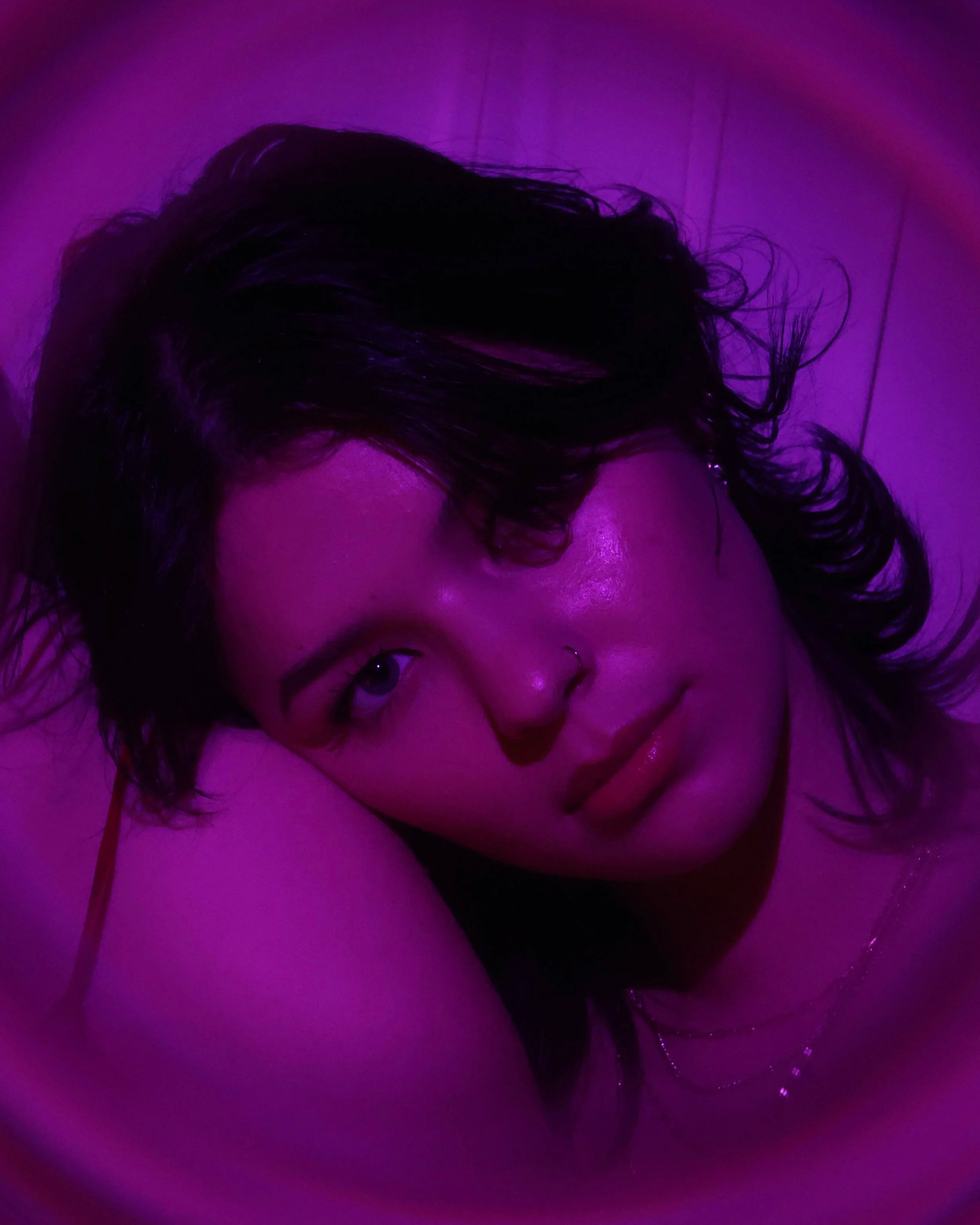 Close-up of a person's face illuminated with purple light, showing dark, curly hair and a nose piercing.