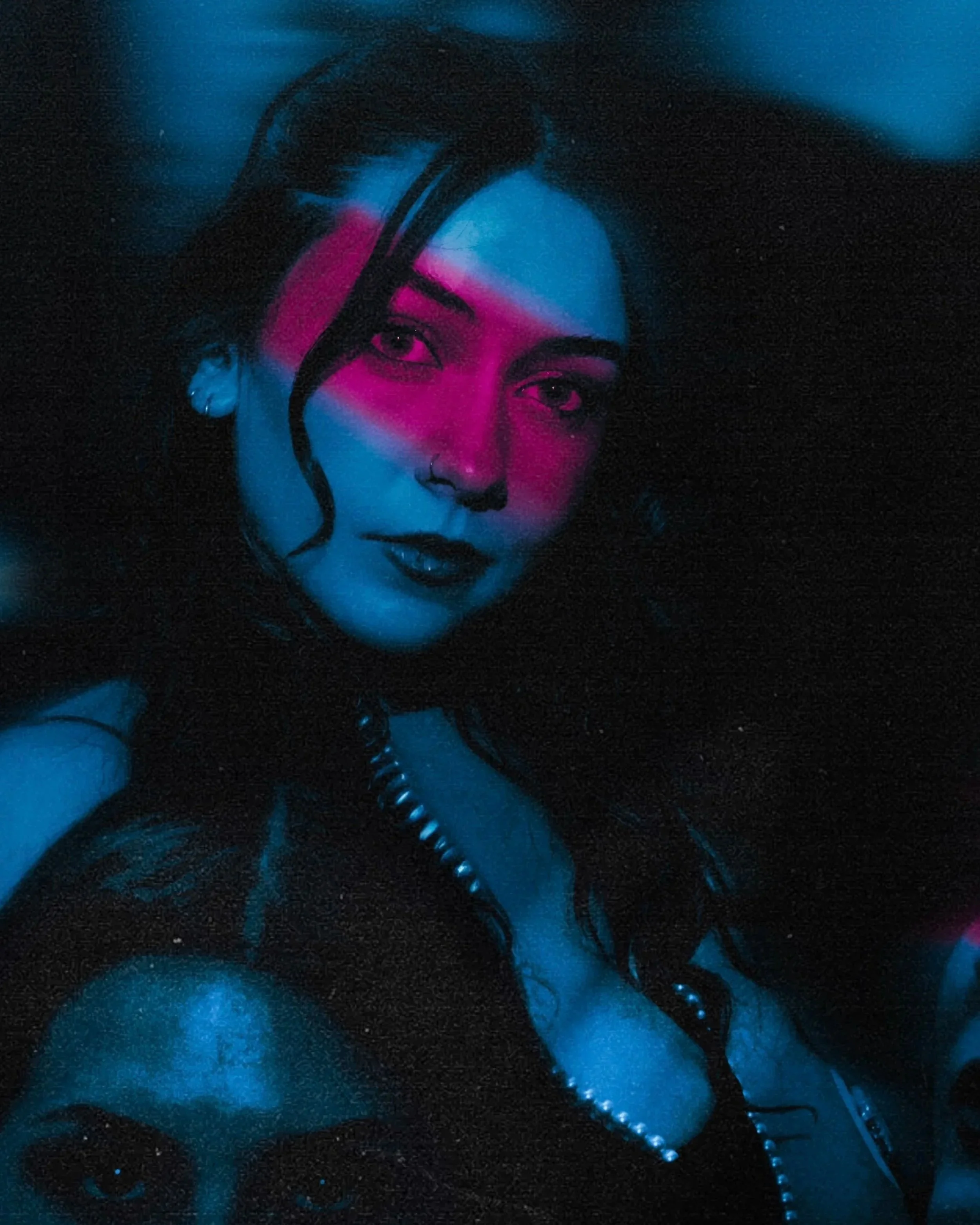 A woman with black hair styled in loose curls, wearing earrings and a pearl necklace, has striking eye makeup and a nose ring, and she is illuminated by blue and pink neon light.
