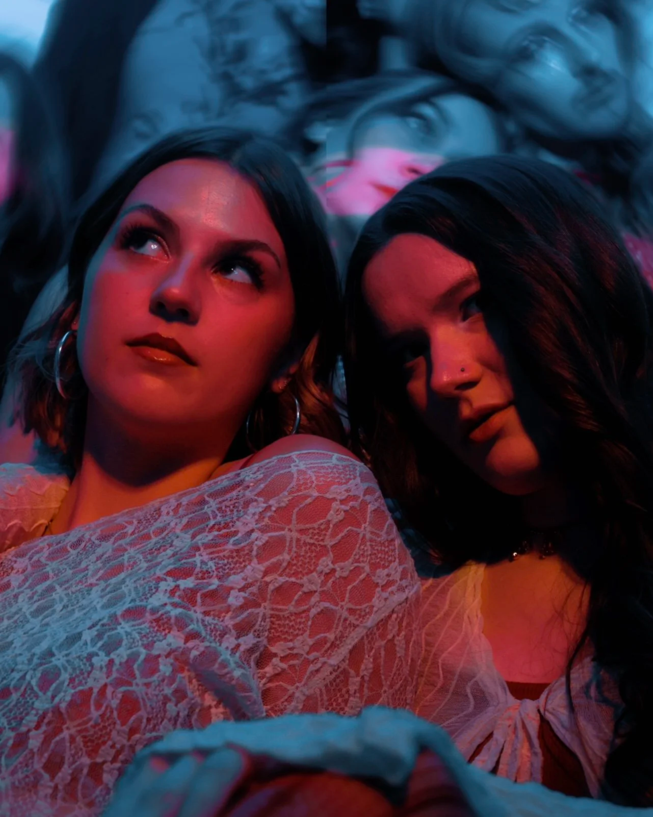 Two women with dark hair lying down together, illuminated by pink and blue lighting, wearing white lace clothing.