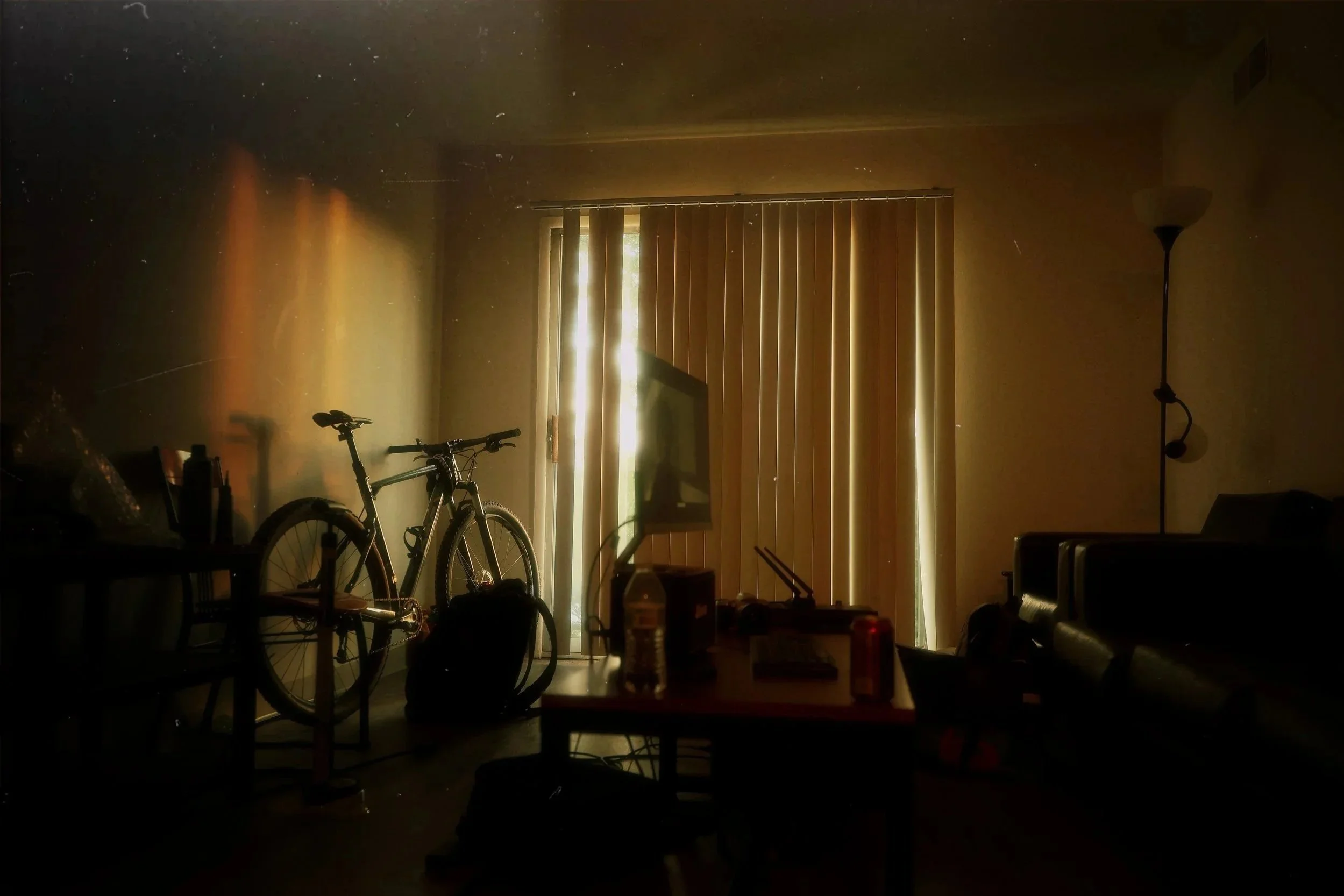 Sunlight filtering through vertical blinds into a dim living room, with a bicycle leaning against the wall, and various furniture and objects scattered around.