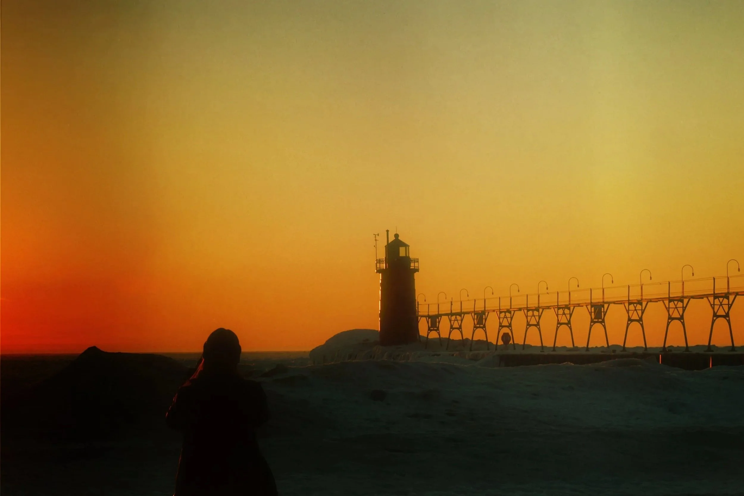 A person sitting on rocks near a beach during sunset, with a lighthouse and a pier in the background.