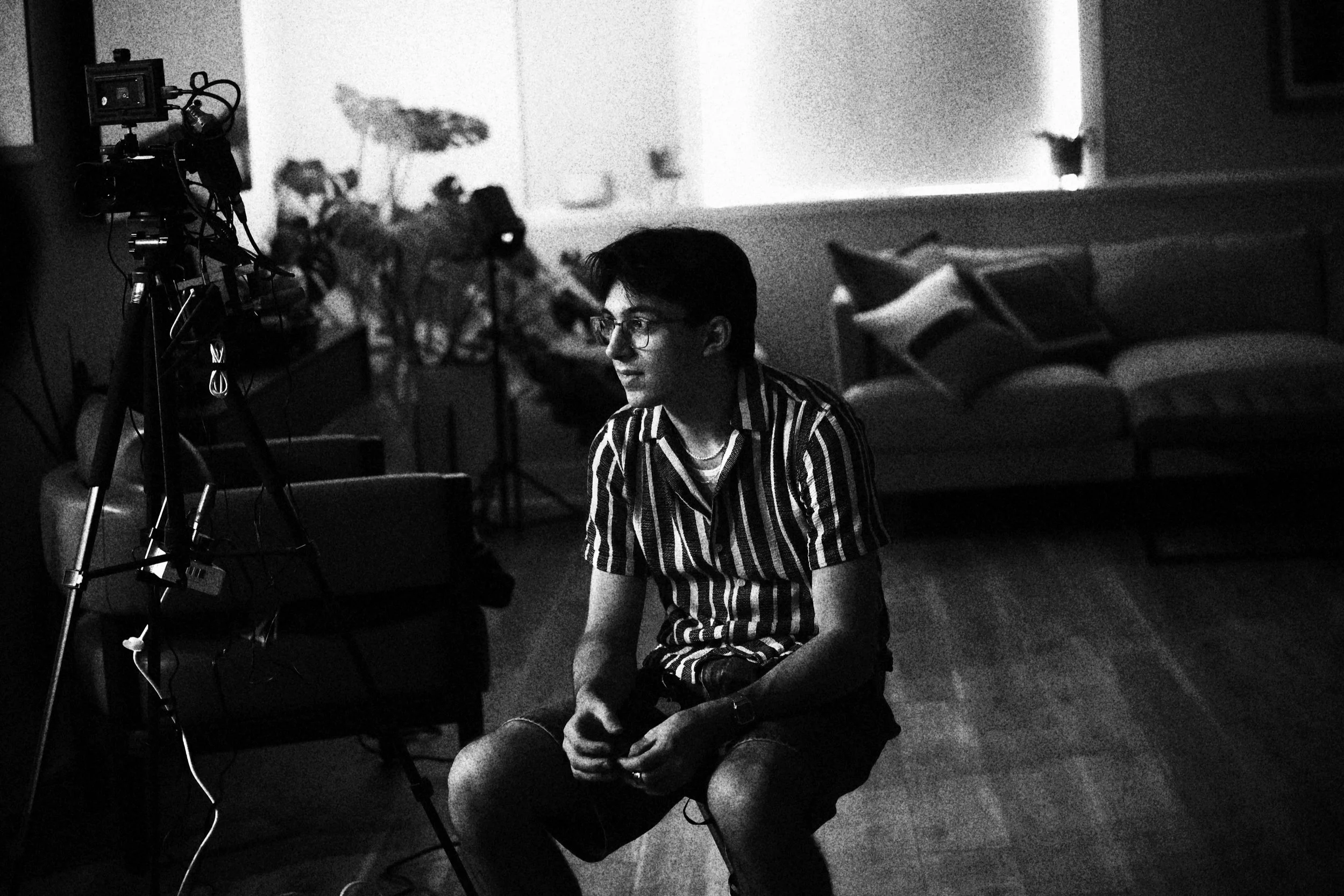 A black and white photo of a person with short dark hair and glasses sitting on the floor in a room, surrounded by electronic equipment, with a sofa and pillows in the background.