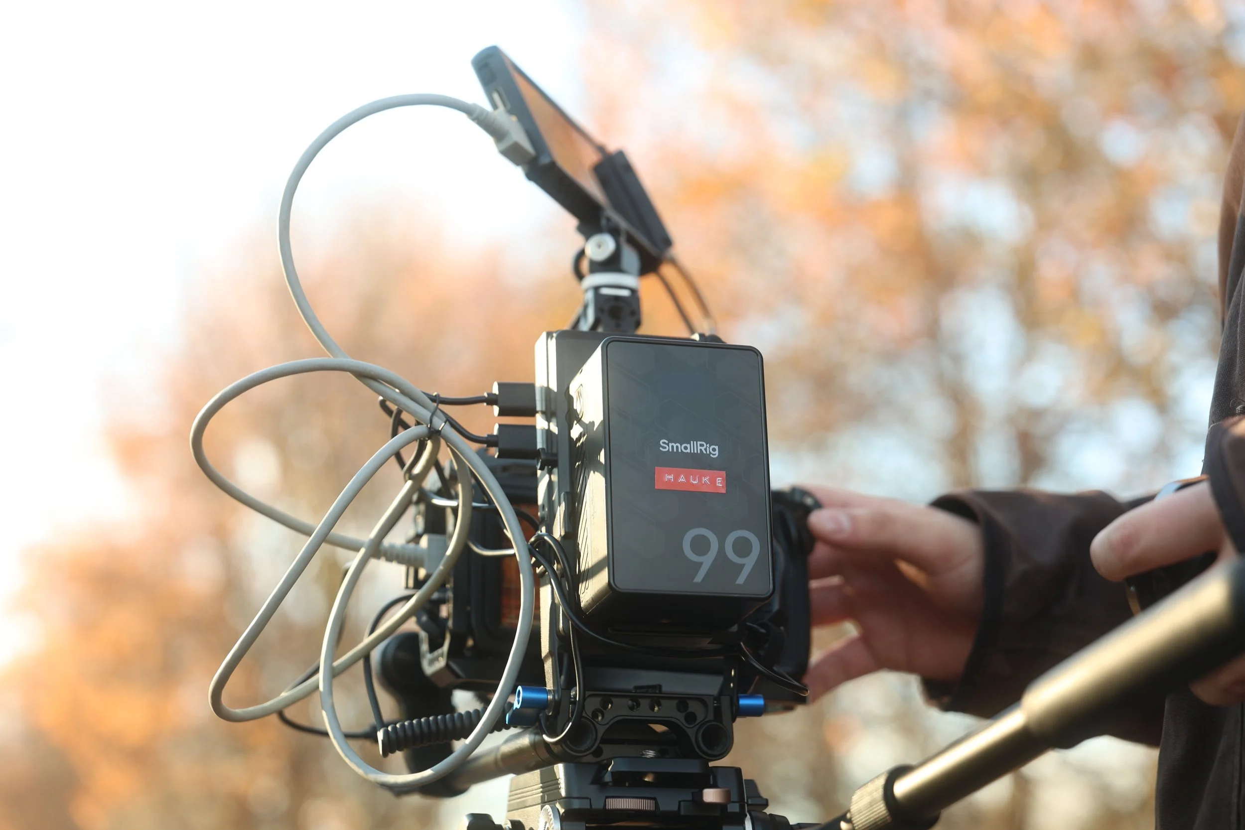 Close-up of a camera mounted on a bike handlebar, with a person's hand adjusting controls, and a blurred autumn background.
