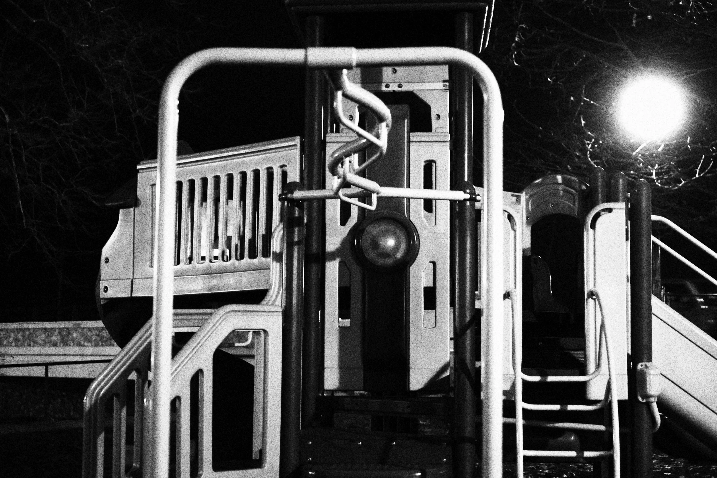 Nighttime black and white photo of a playground structure with slides and climbing bars, illuminated by a bright light in the background.