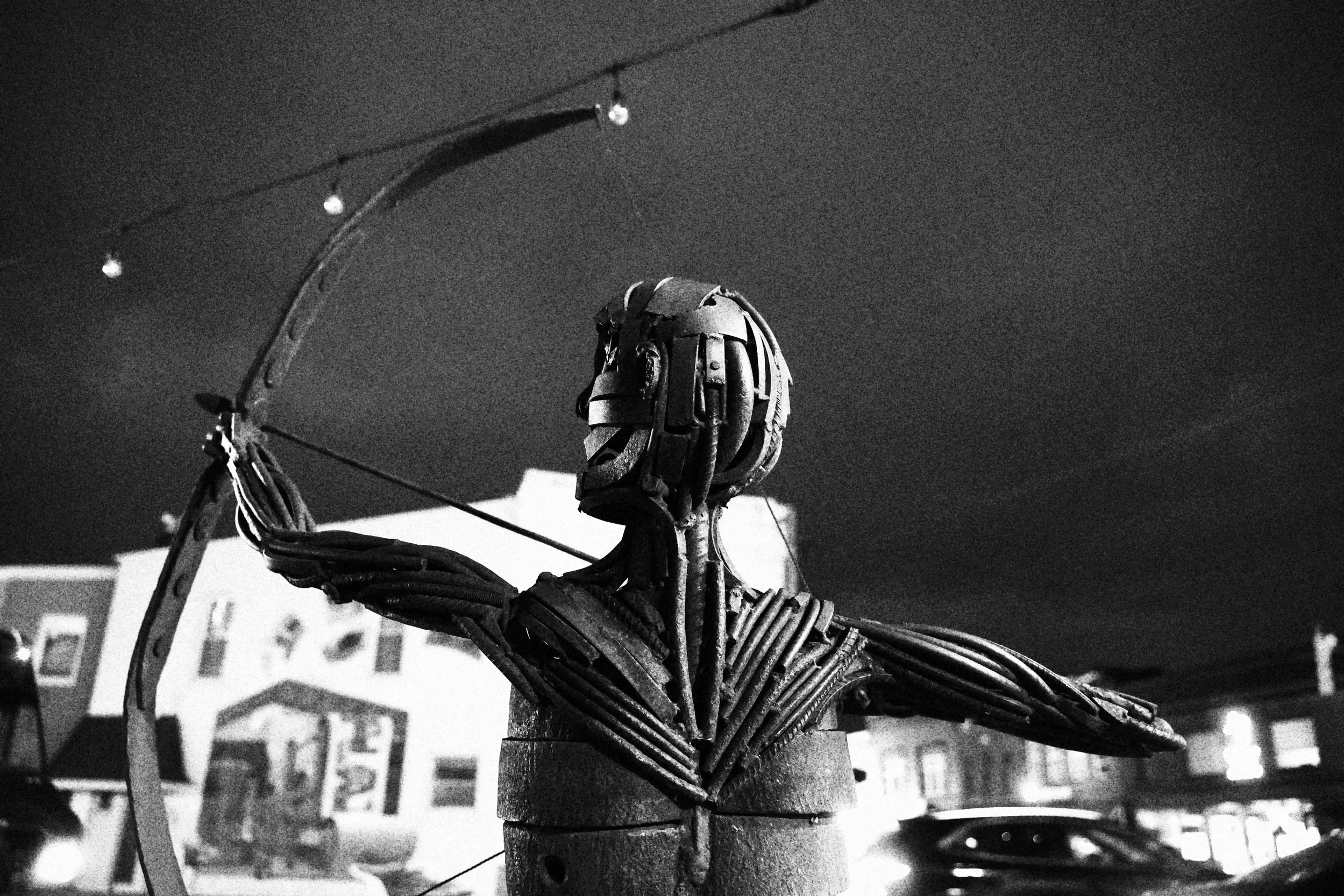 A black and white image of a metal robot sculpture made from various mechanical parts, standing outdoors under string lights with blurry buildings in the background.