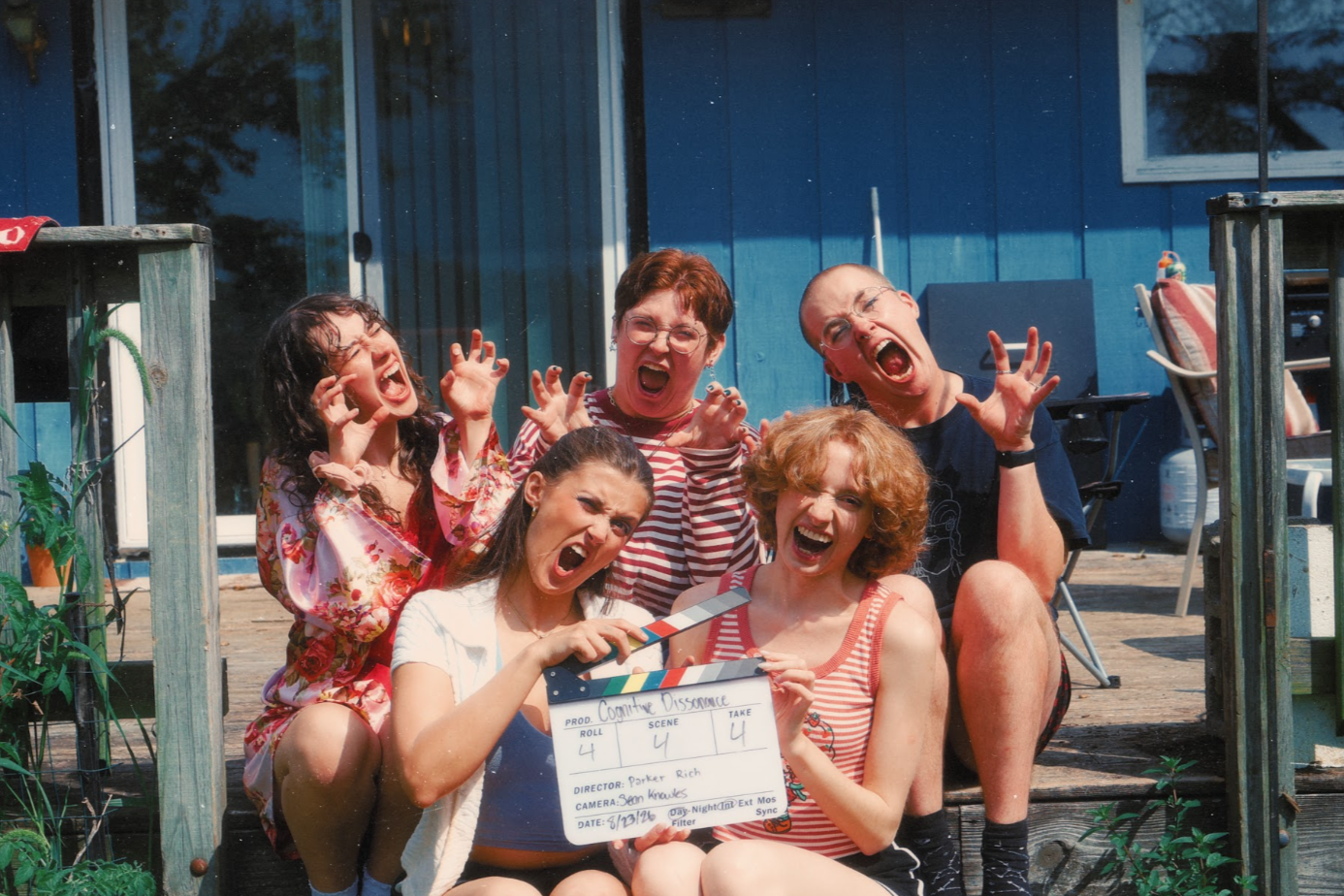 Group of six people making funny faces, one holding a clapperboard, on a wooden deck outside a blue house.