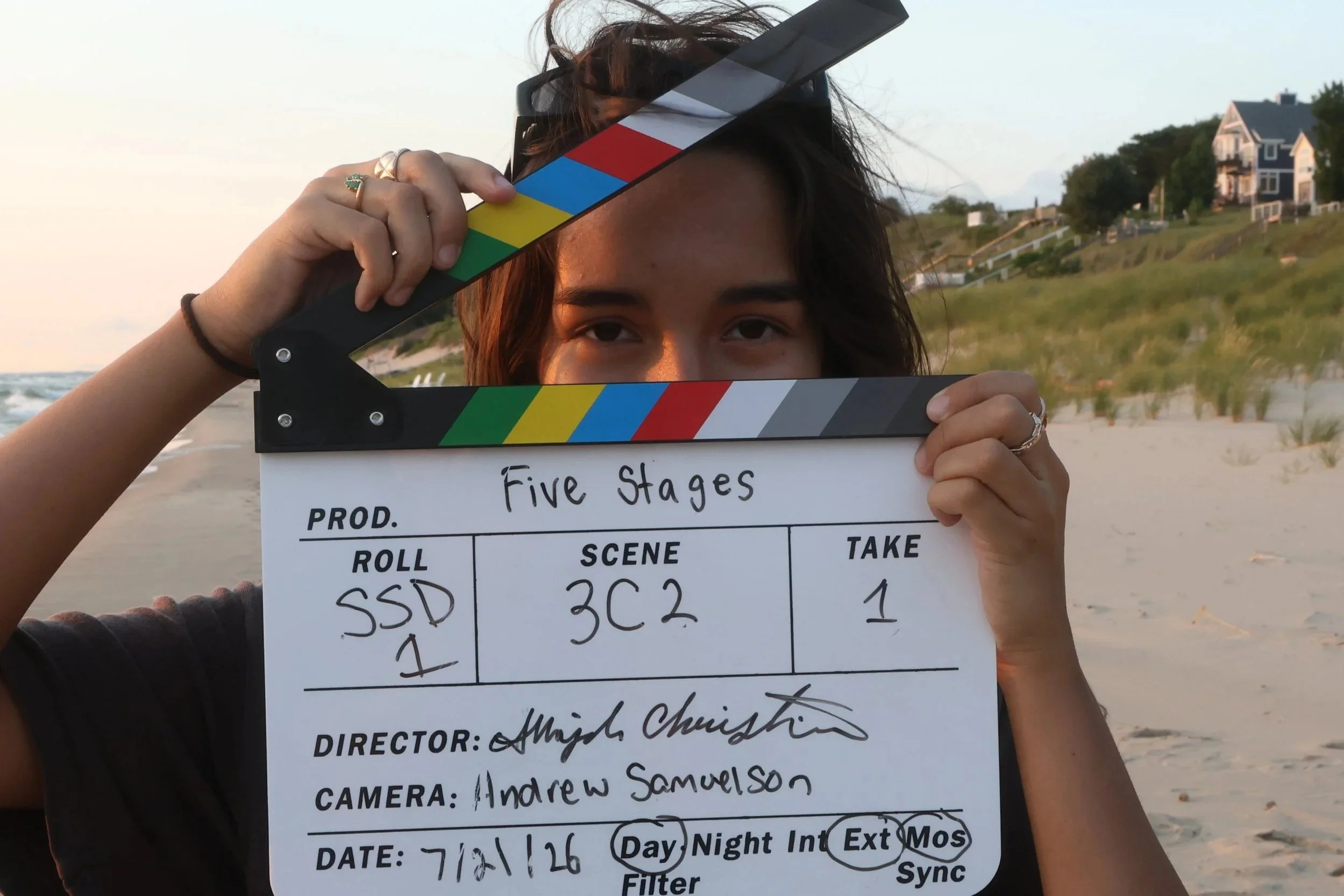 A person holding a film slate with notes, standing on a beach with houses on a hillside in the background at sunset.