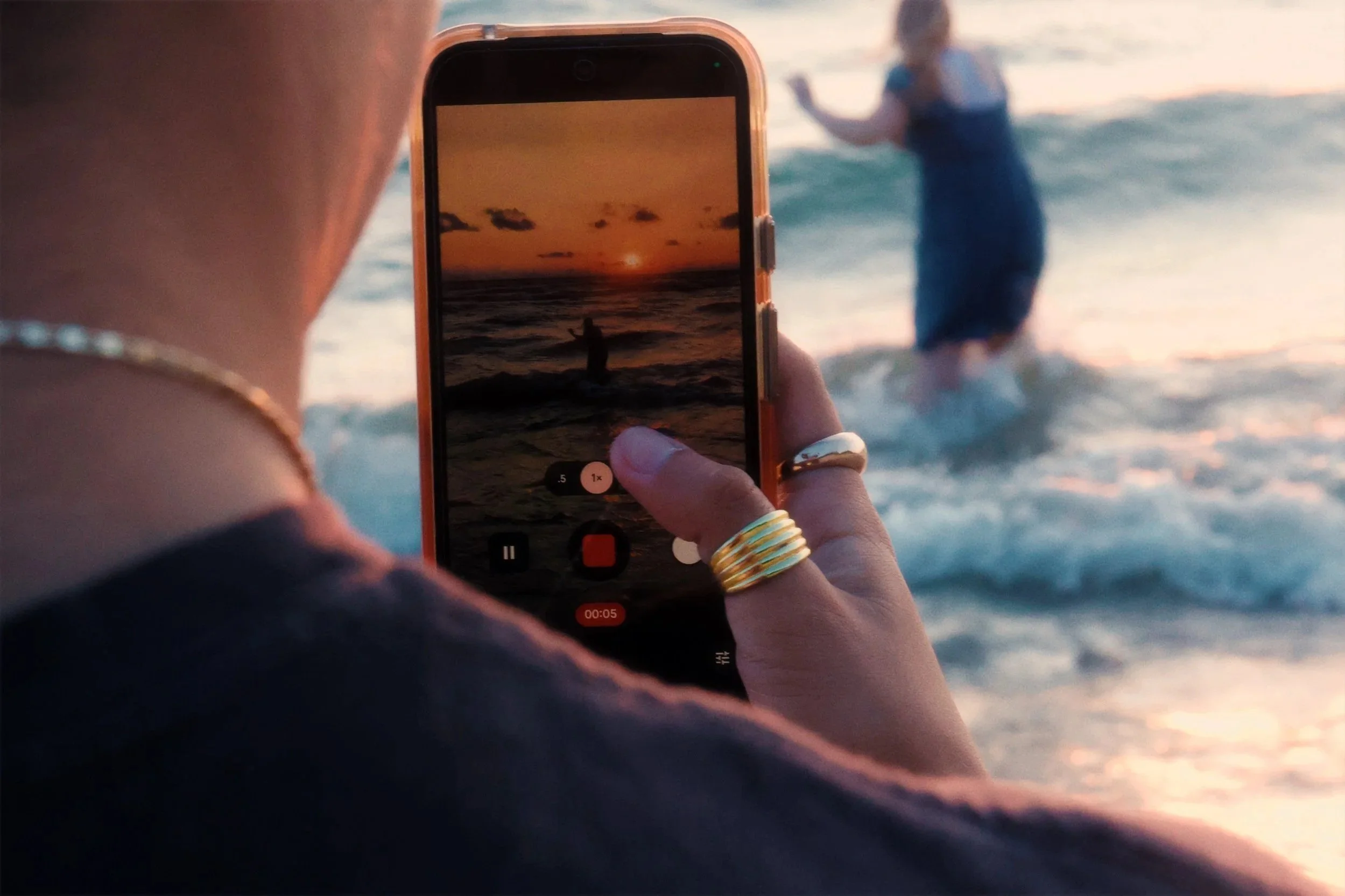 Person taking a photo of a child playing in the ocean during sunset with a smartphone, close-up of hand with gold rings, blurred background of waves and sky.