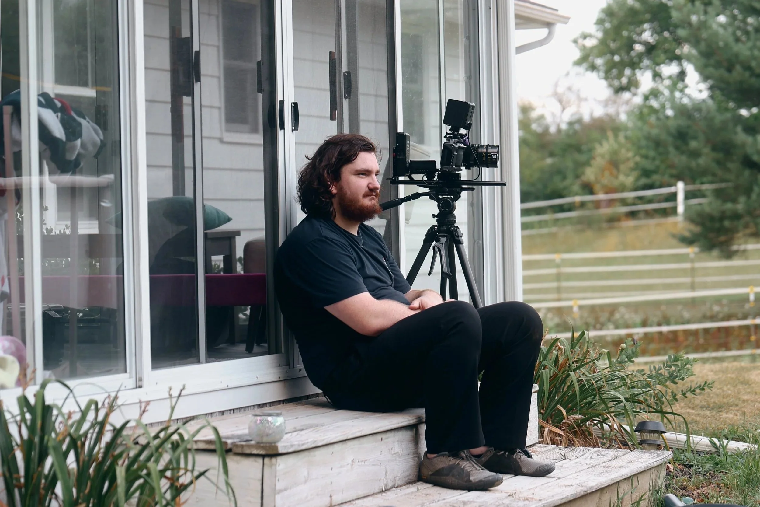 A man with long dark hair and a beard sitting on the porch in front of a glass window, operating a professional camera mounted on a tripod.