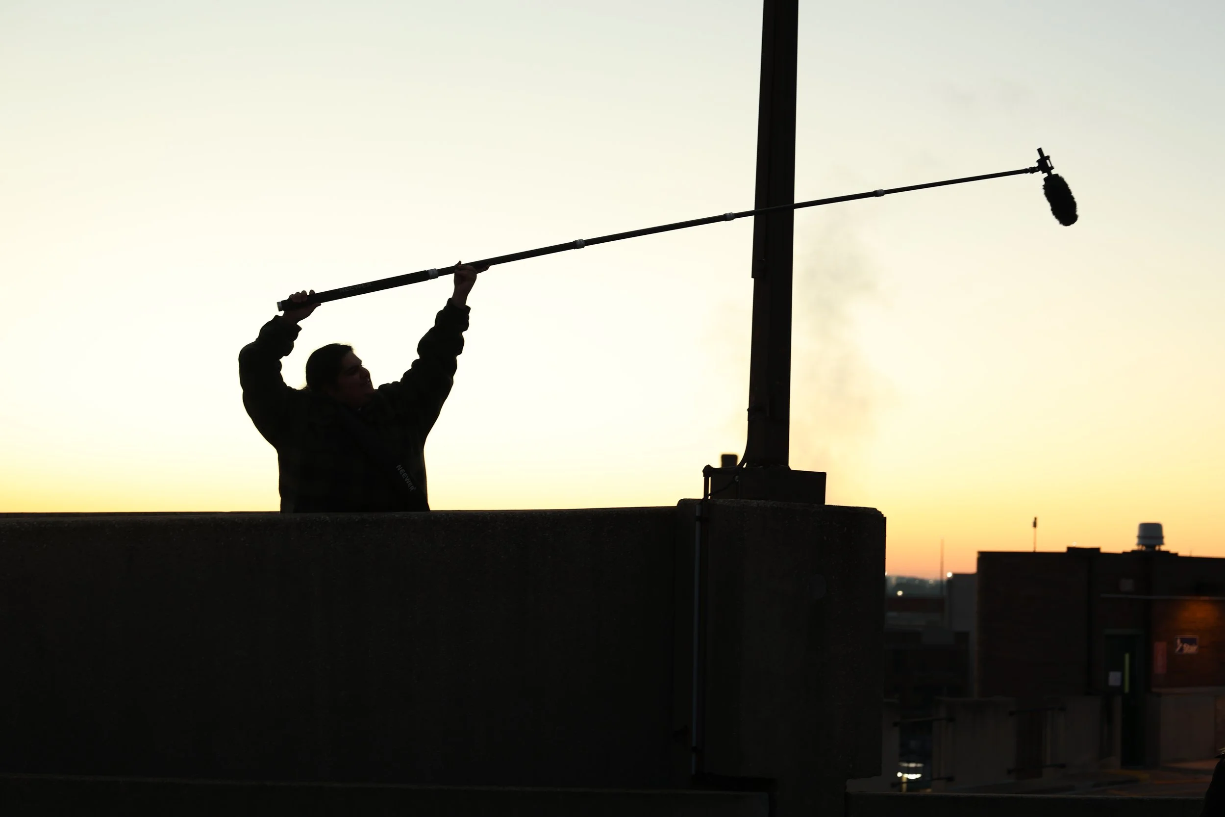 A person holding a boom microphone on a rooftop during sunset, with city buildings in the background.