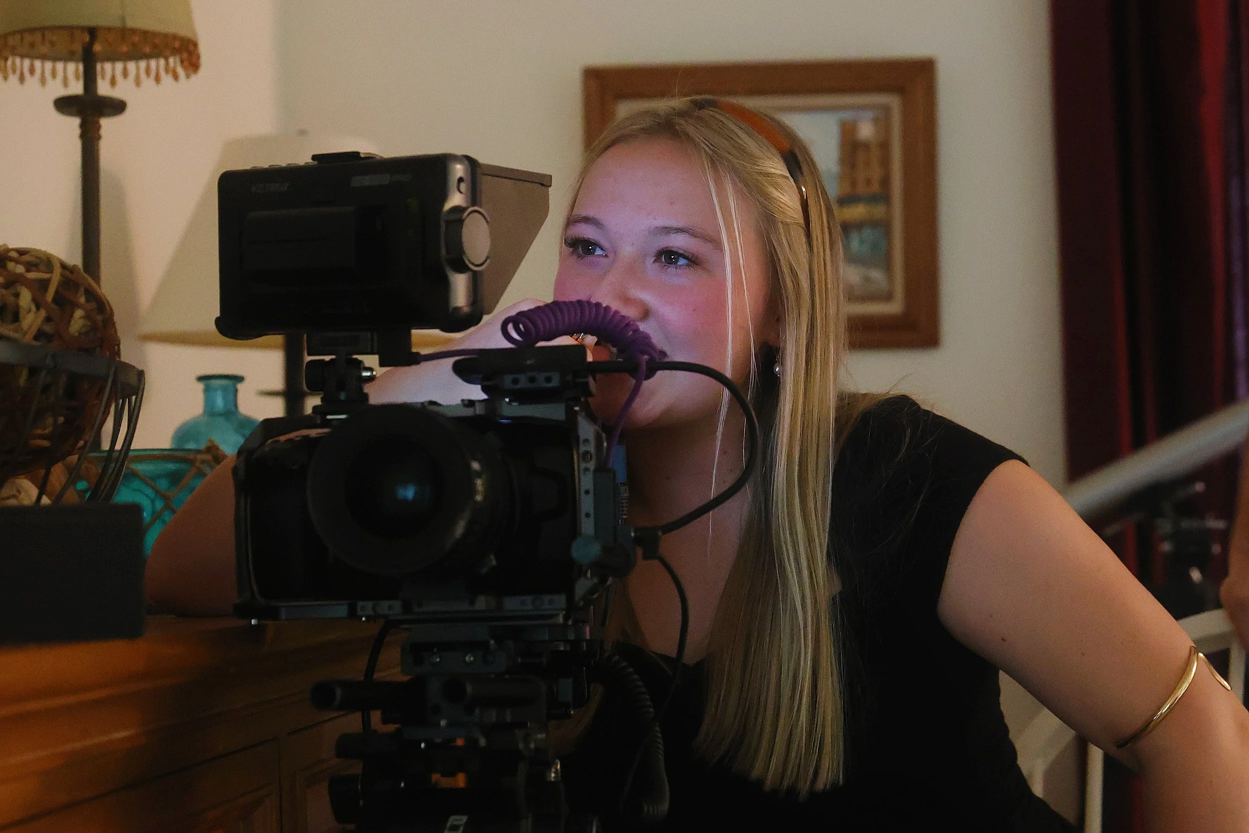 Young woman with long blonde hair, wearing headphones, looking through a camera mounted on a tripod, in a room with a wooden desk, decorative lamp, and framed pictures on the wall.