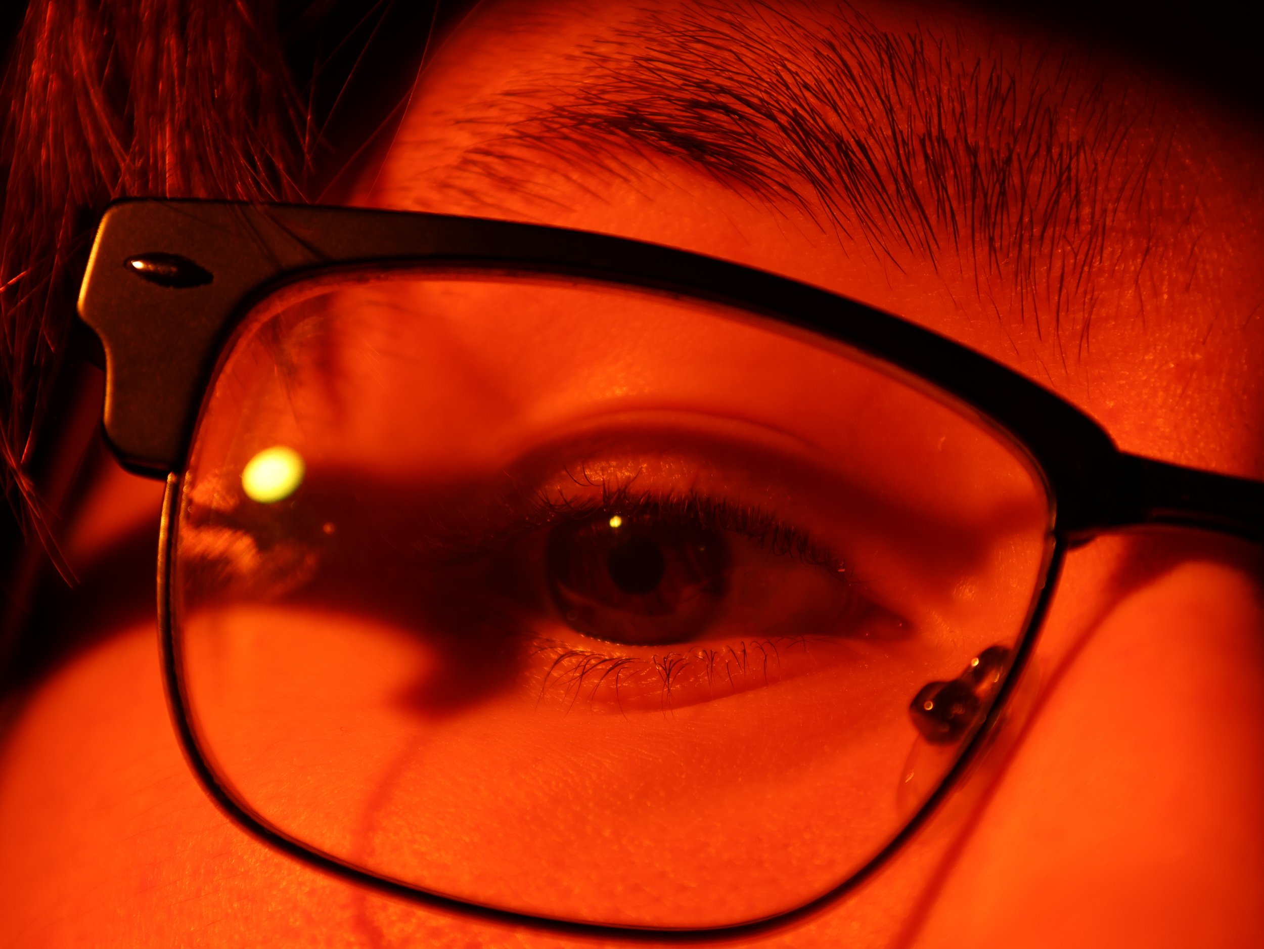 Close-up photo of a person's eye wearing glasses, with the reflection of a bright light, and detailed eyelashes and skin visible.
