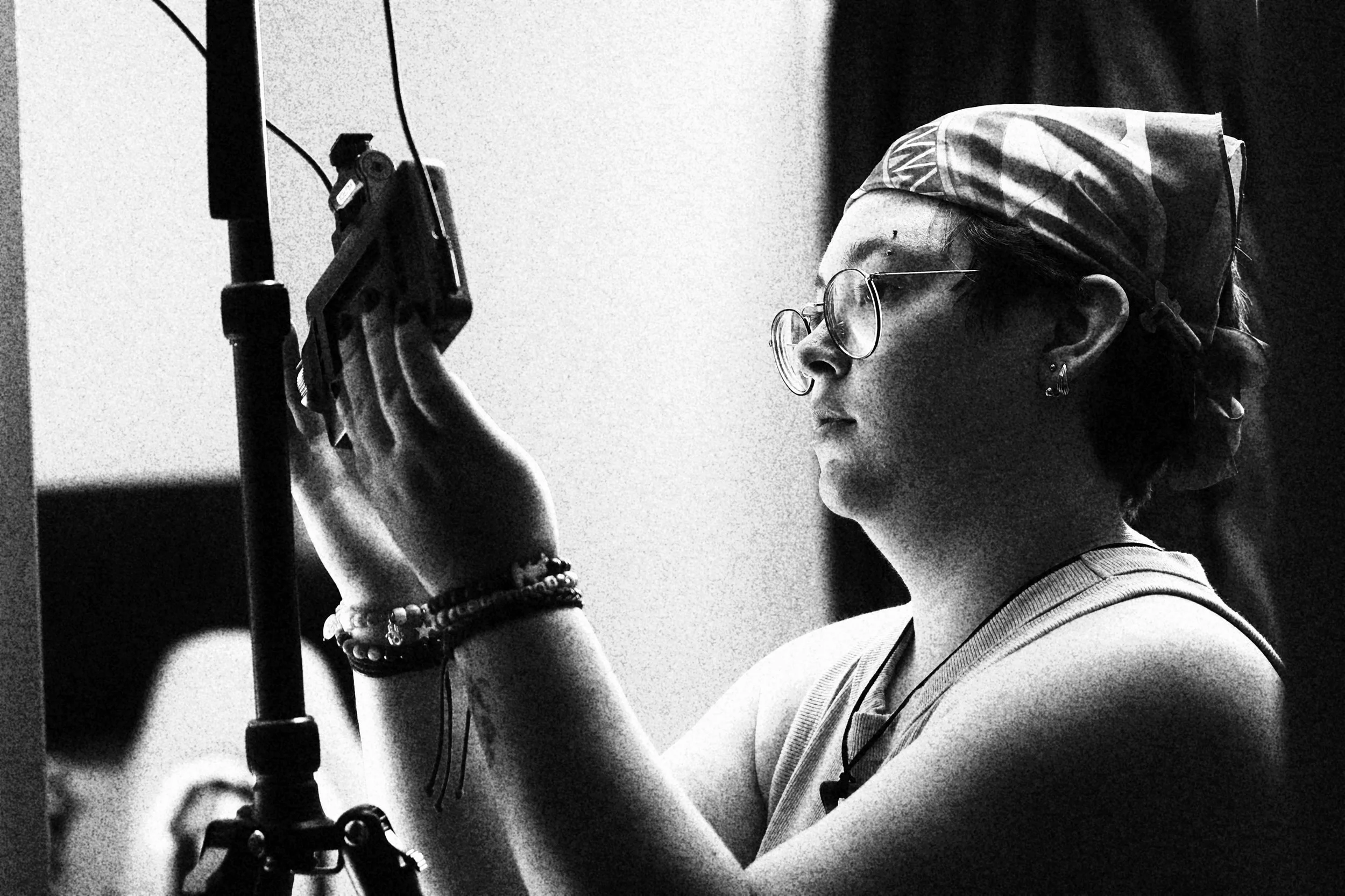 A woman wearing glasses, a patterned headscarf, and multiple bracelets, looking through a viewfinder or camera, in black and white.