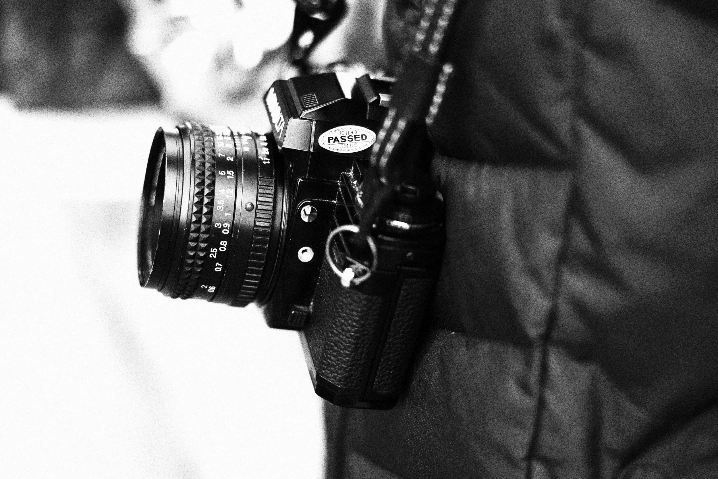 A black and white photograph of a vintage film camera placed on a dark surface, with a focus on the camera's lens and body, featuring an 'APPROVED' sticker on it.