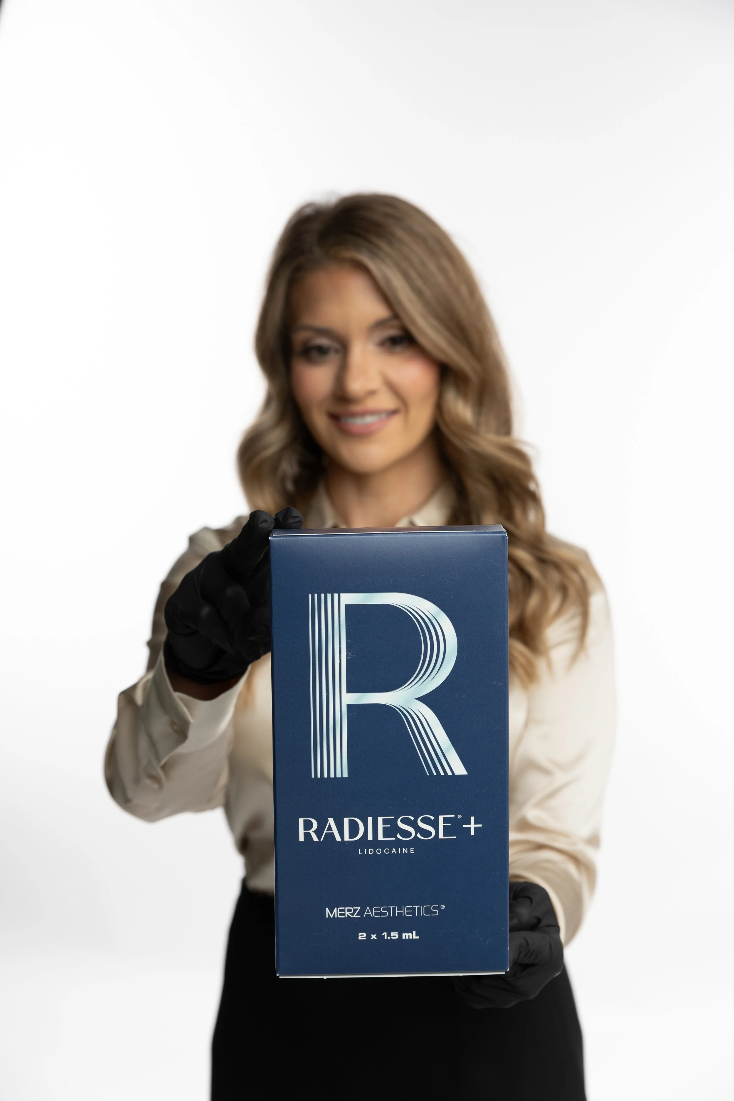 Woman in a beige blouse and black gloves holding a box of Radiesse+, a dermal filler product.