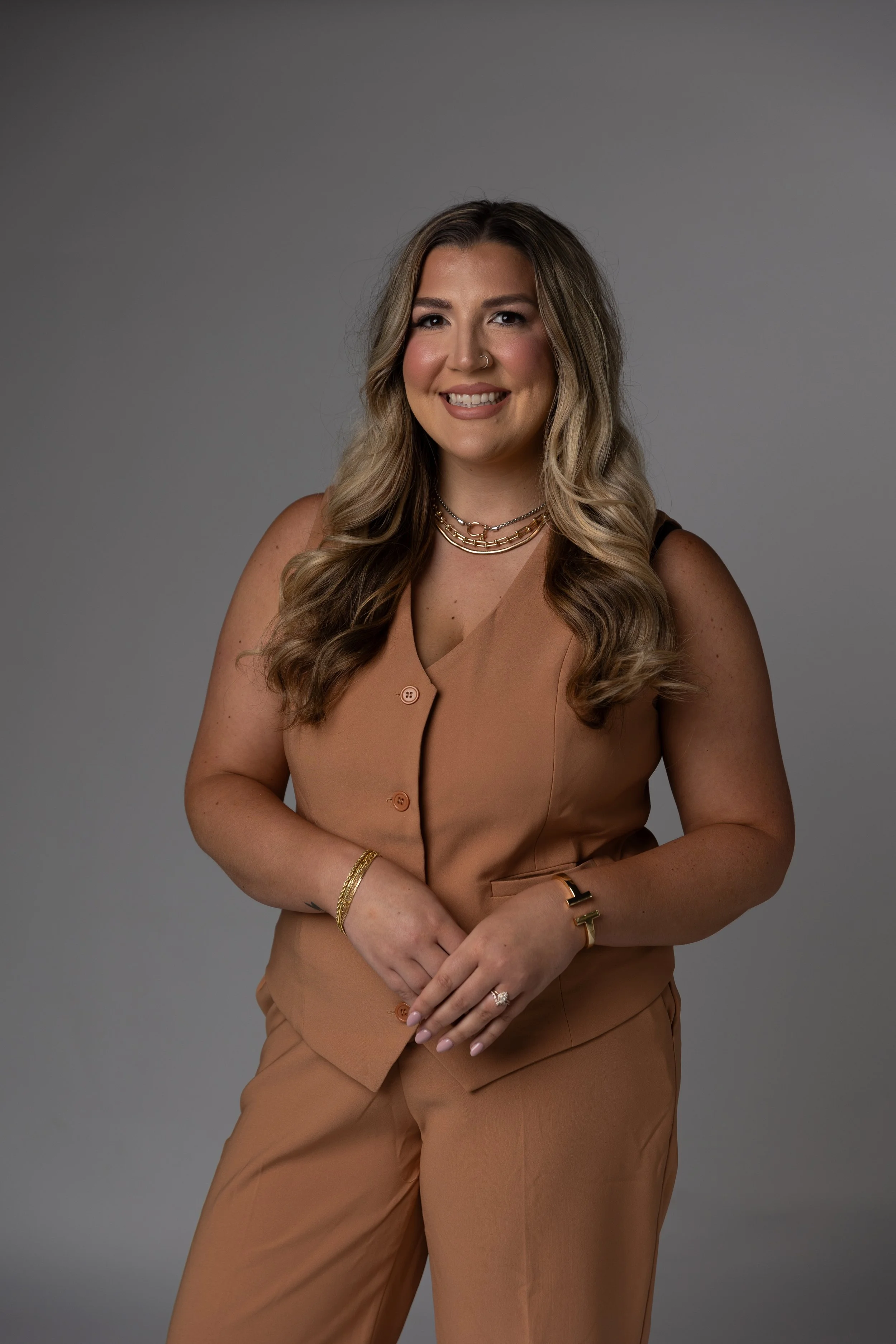 A woman with long wavy blonde hair smiling while standing against a gray background, wearing a tan sleeveless blazer and matching pants with gold jewelry.