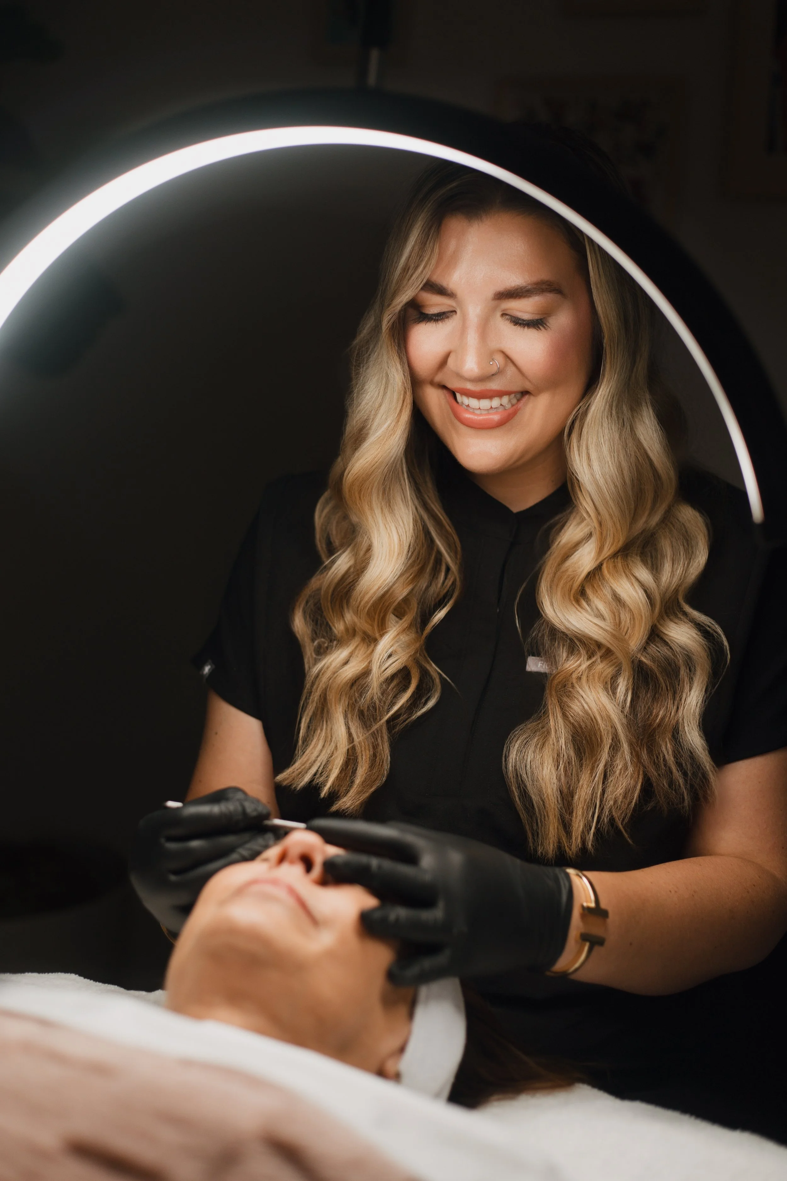 A woman with long, wavy blonde hair smiling while performing a cosmetic procedure on a patient lying down, with her face covered by a cloth, using medical tools. The scene is illuminated by a bright ring light.