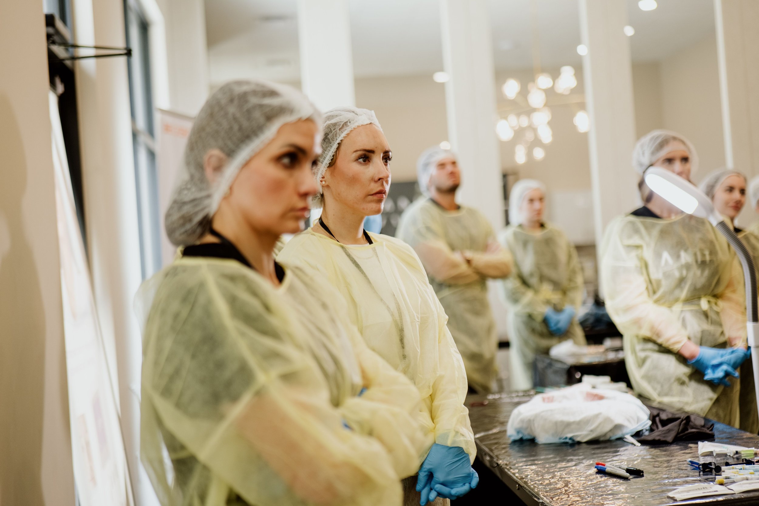 Group of people in yellow protective gowns, hair nets, and gloves attending a medical training or seminar in a well-lit room.