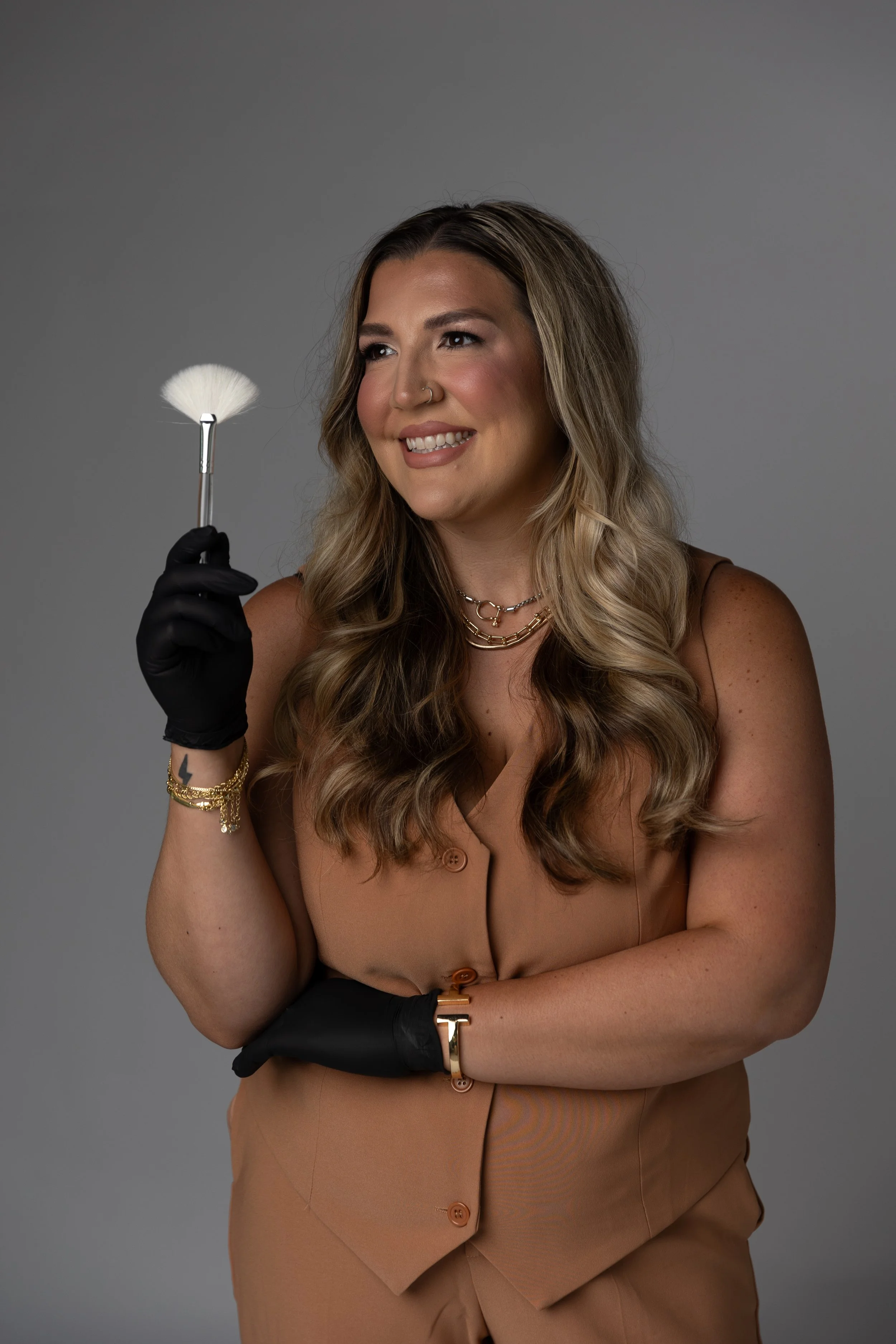 A woman with wavy brown hair wearing a tan outfit and jewelry, holding a makeup brush, smiling, against a gray background.