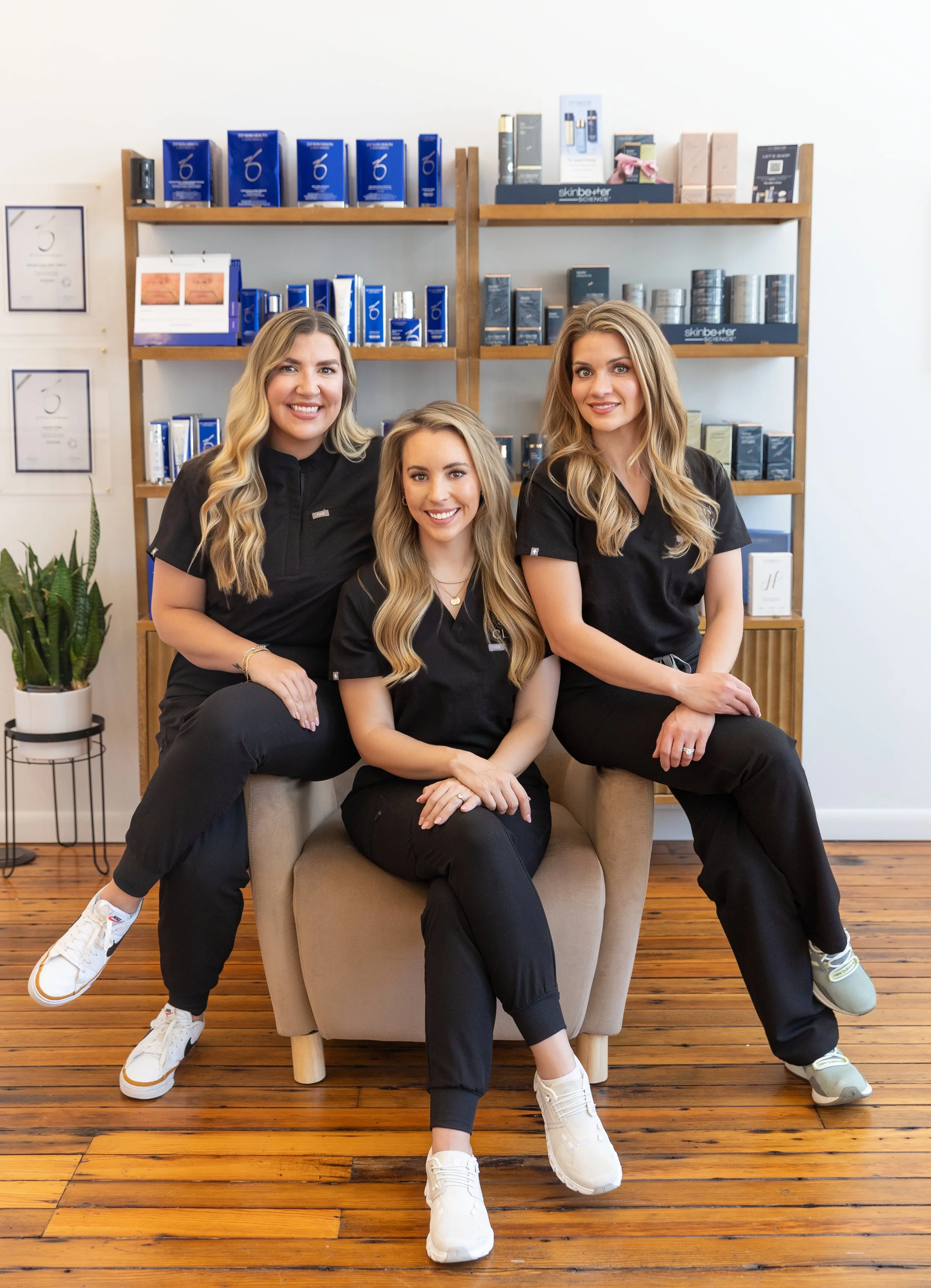 Three women sitting and standing on a beige chair in a skincare or salon store, smiling, wearing black uniforms, with shelves of skincare products behind them and a potted plant to the side. Wooden flooring is visible.
