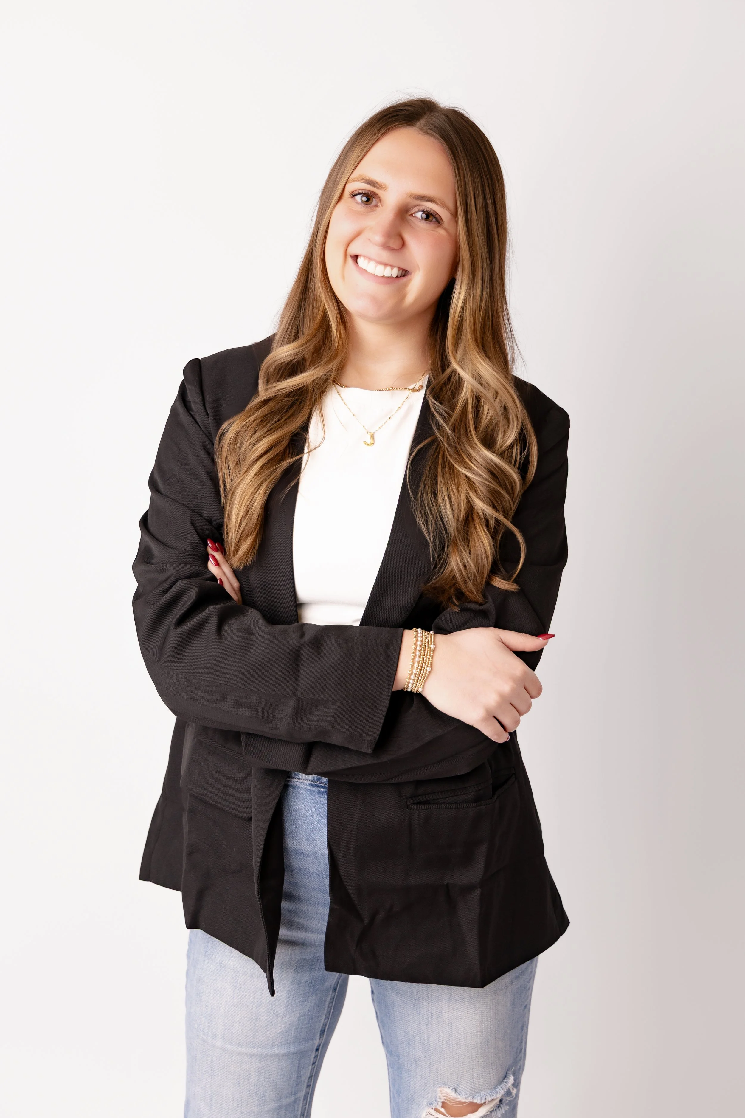 Portrait of a young woman with long wavy hair, wearing a black blazer, white top, light blue ripped jeans, gold jewelry, and smiling with arms crossed against a plain white background.