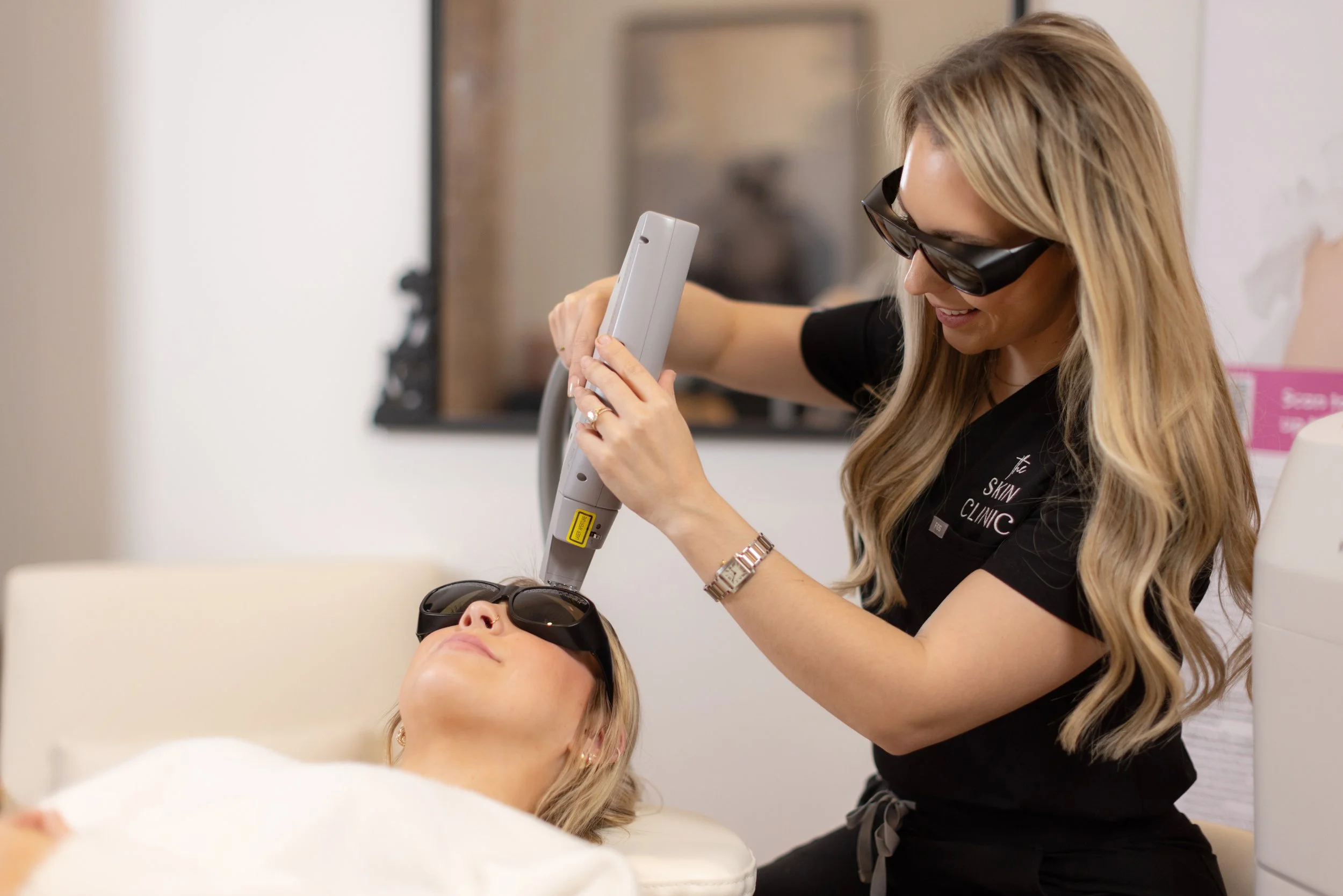 A woman lying on a treatment bed receiving laser skin treatment from a technician in a clinic, both wearing protective goggles.