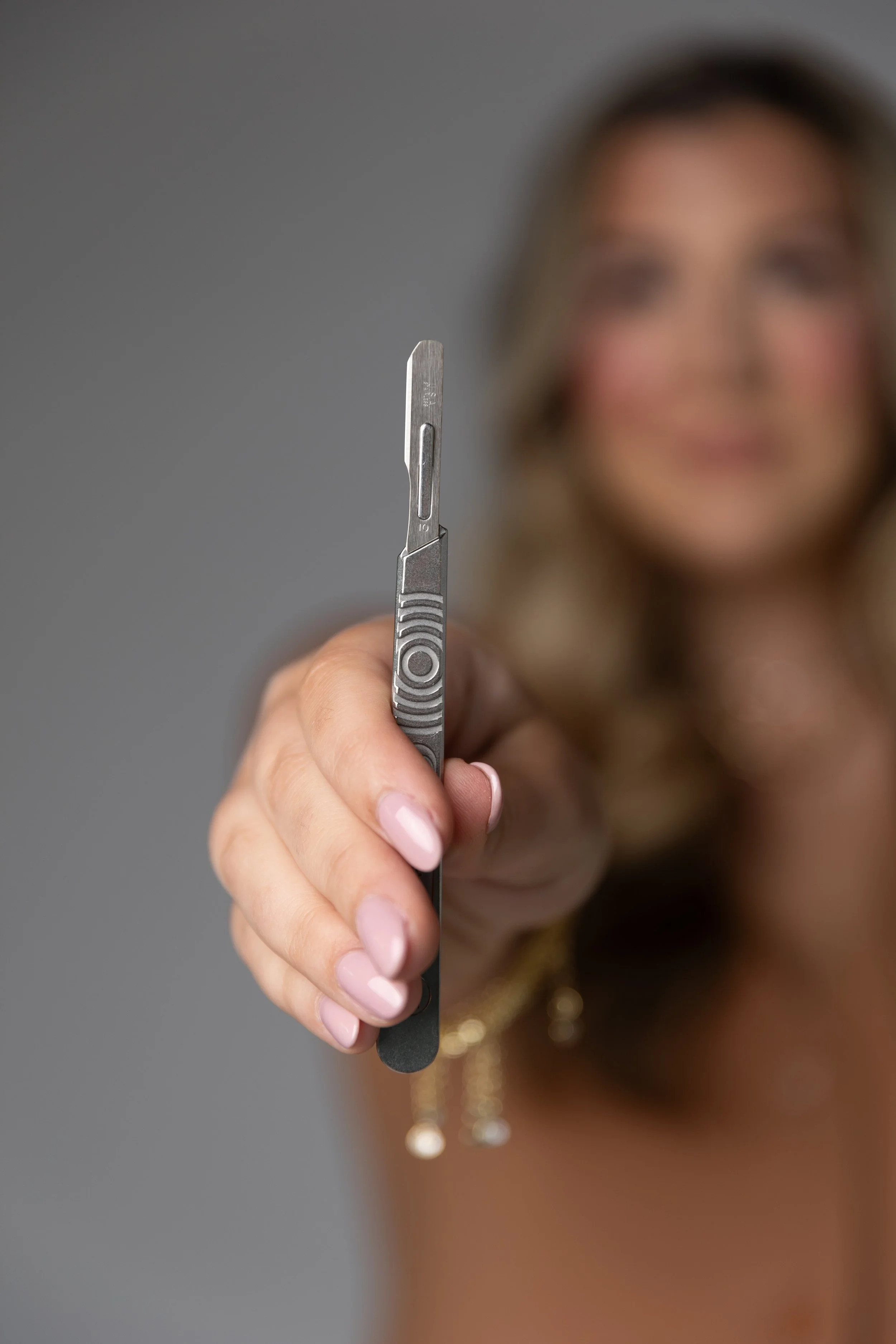 Close-up of a woman with blond hair and light skin holding a small precision scalpel toward the camera, with a blurred face in the background.