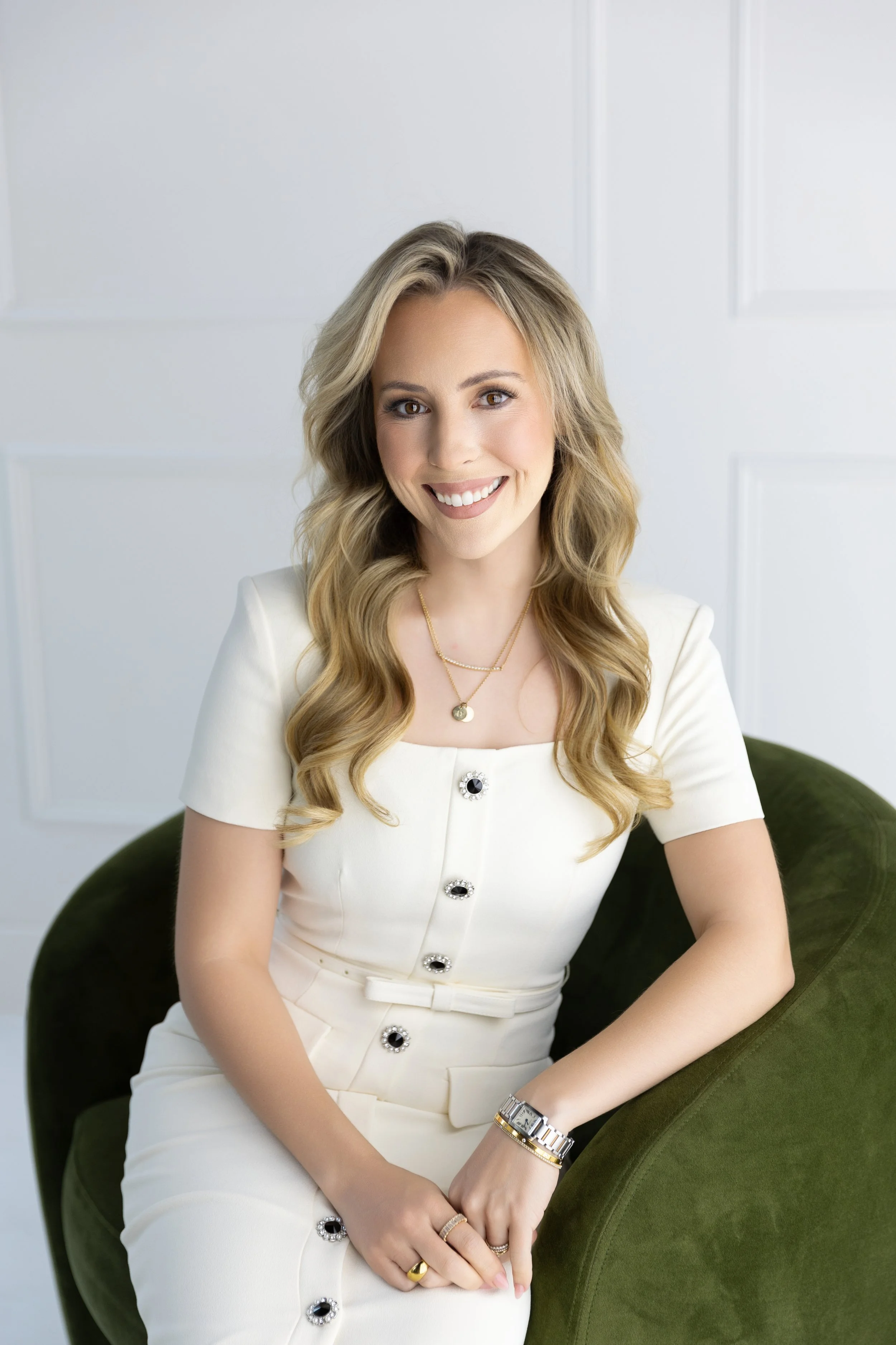 A smiling woman with wavy blonde hair in a white dress with black and silver buttons, sitting in a green velvet armchair against a white wall.