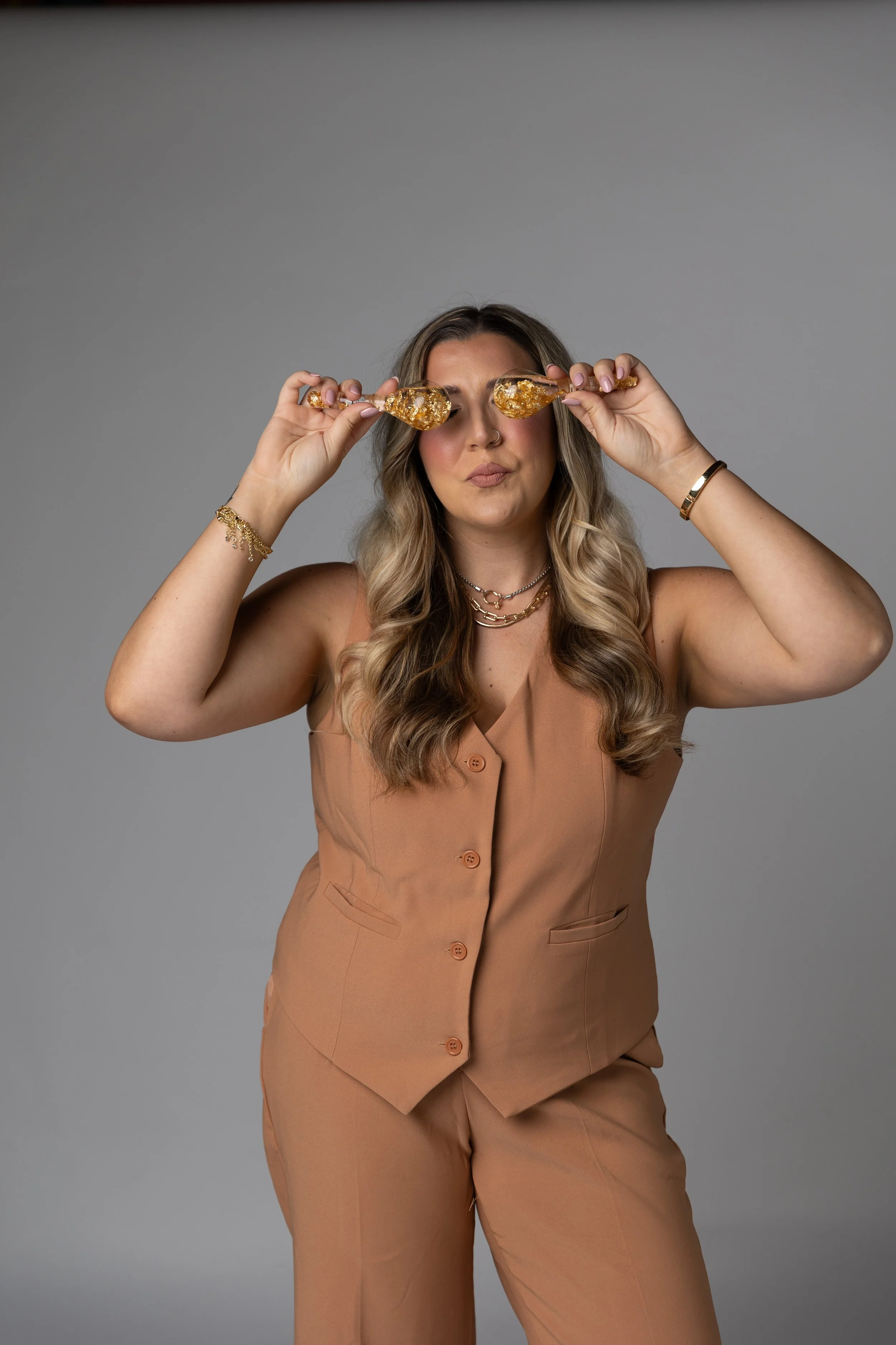 A woman with wavy blonde hair is holding two gold glittery champagne glasses near her eyes, making a playful expression. She is wearing a tan sleeveless vest and matching pants, accessorized with layered necklaces and bracelets, standing against a plain gray background.